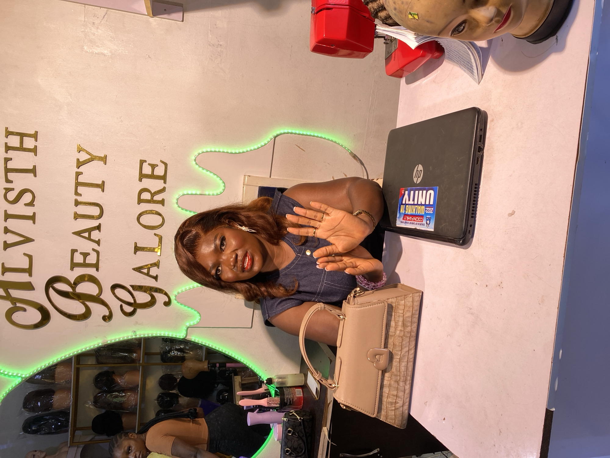 In the city of Bukuru, Nigeria, a woman sits behind a counter in what appears to be a beauty salon. The wall behind her is adorned with golden lettering spelling "ALVISTH BEAUTY GALORE," outlined by a vibrant green neon light. The woman is wearing a denim-like top and has her hands raised in front of her, palms facing outward. On the counter in front of her sits a tan handbag with a heart-shaped clasp, a black laptop with stickers, and to the right, a red sewing machine and a mannequin head with stylized hair. To the left of the woman, a mirror reflects shelves stocked with various wigs. Another woman, wearing an orange top and dark leggings, is visible in the reflection of the mirror, looking towards the camera. The overall scene suggests a commercial establishment focused on beauty products and services.