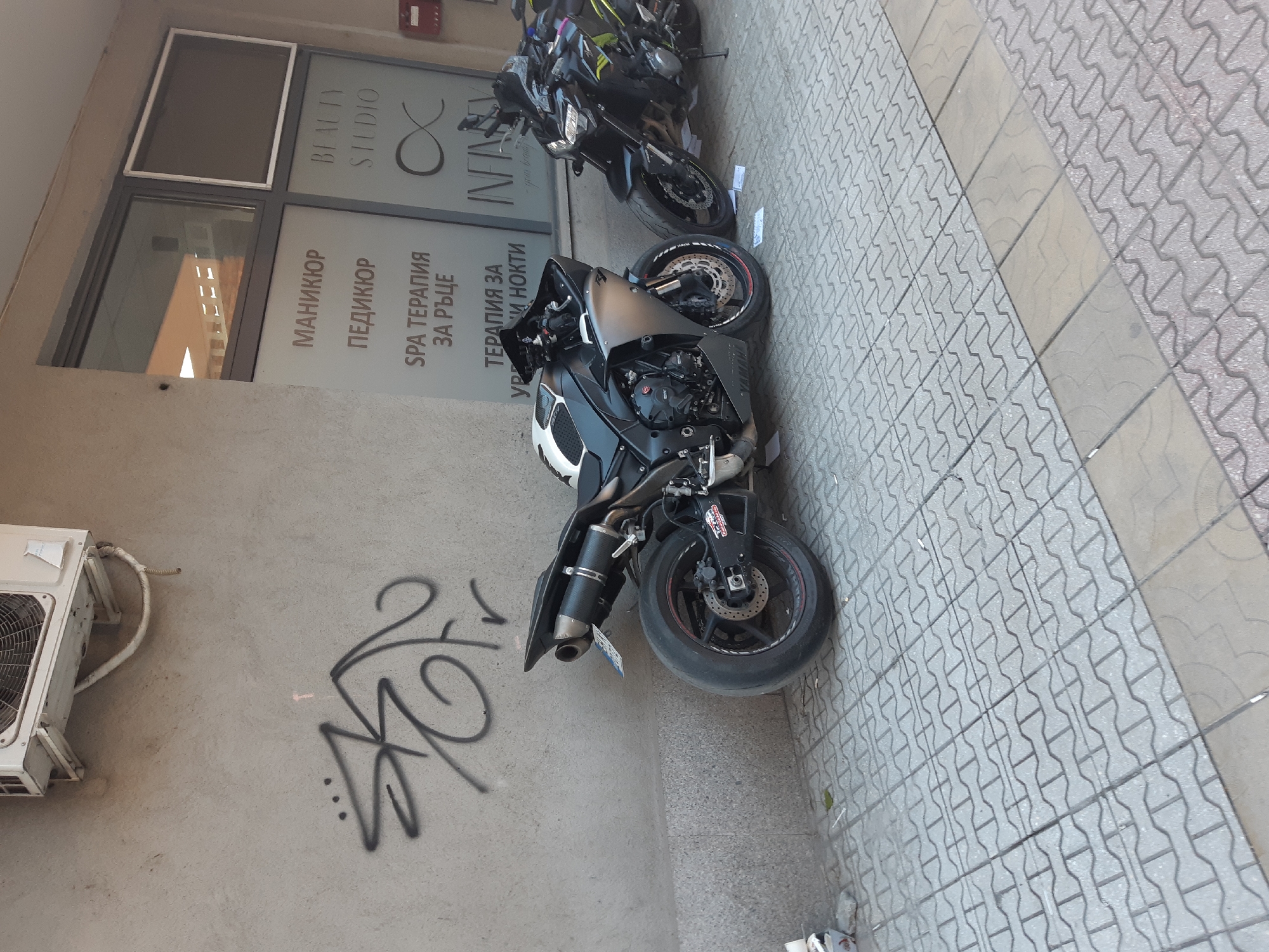 The image depicts two motorcycles parked on a paved sidewalk next to a building in Plovdiv, Bulgaria. The building has a sign indicating a beauty studio called "Infinity," with services listed in Bulgarian, including manicure, pedicure, and spa therapy. Graffiti is visible on the wall beside the motorcycles and an air conditioning unit is mounted on the wall.