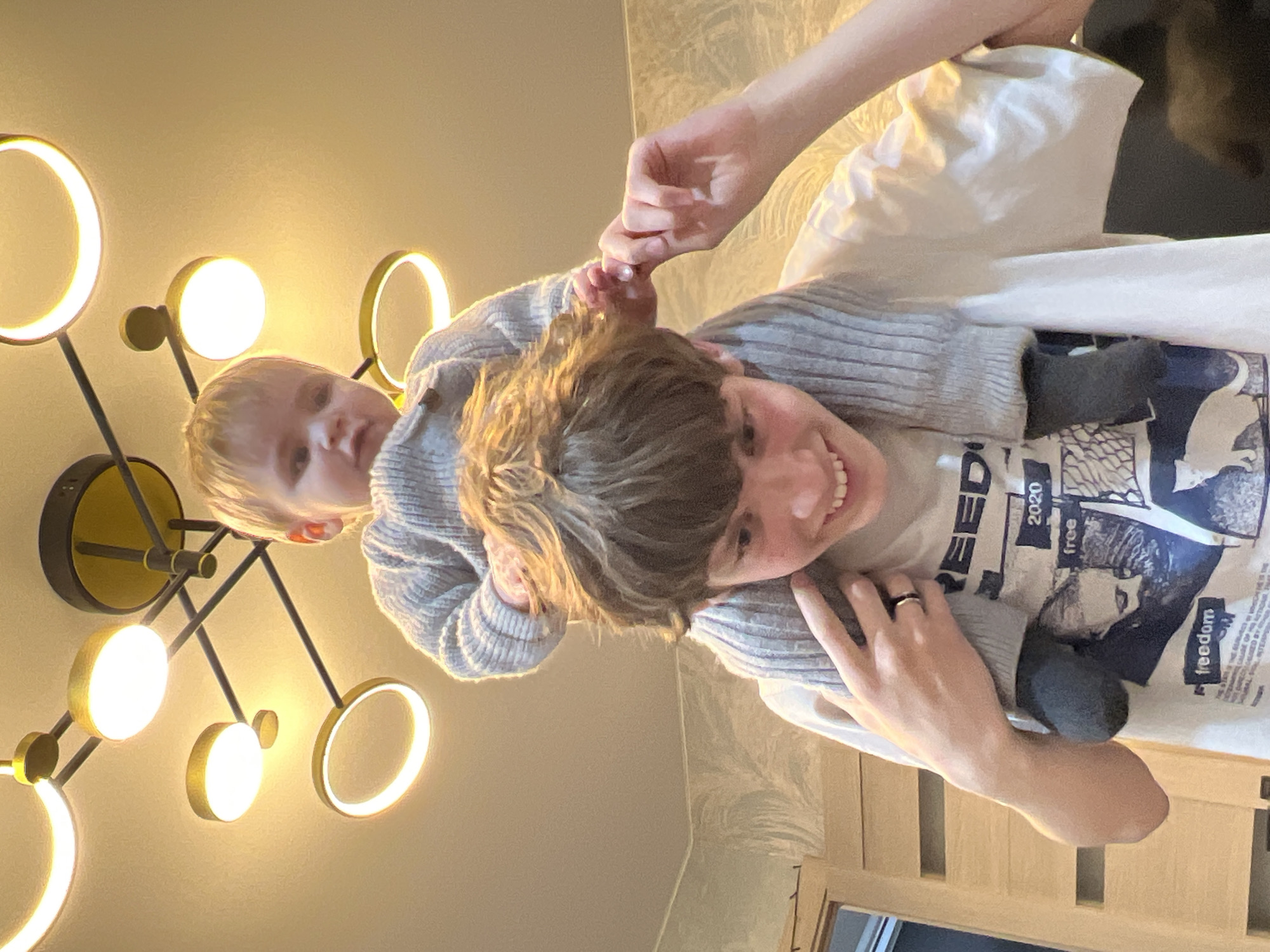 An adult and a young child are captured in a joyful, candid moment indoors. The adult, with medium-brown hair and a wide smile, is carrying the child on their shoulders or back. The child, with light blonde hair, looks directly at the camera with an inquisitive expression, holding onto the adult's hair. Both are wearing matching or similar grey ribbed sweaters, suggesting comfort wear. The adult's light-colored t-shirt, visible beneath the sweater, features a graphic design with the words "2020", "free", and "freedom". A black ring is visible on the adult's left ring finger.
The scene is illuminated by a striking modern light fixture on the wall/ceiling, which features several circular and ring-shaped glowing elements casting a warm, golden light. The background shows light-colored walls, possibly with a subtle pattern, and a wooden furniture piece, likely a headboard, towards the bottom right. The overall atmosphere conveys warmth, happiness, and a playful interaction, likely taking place in the evening. The location context is Noviy, Russia.