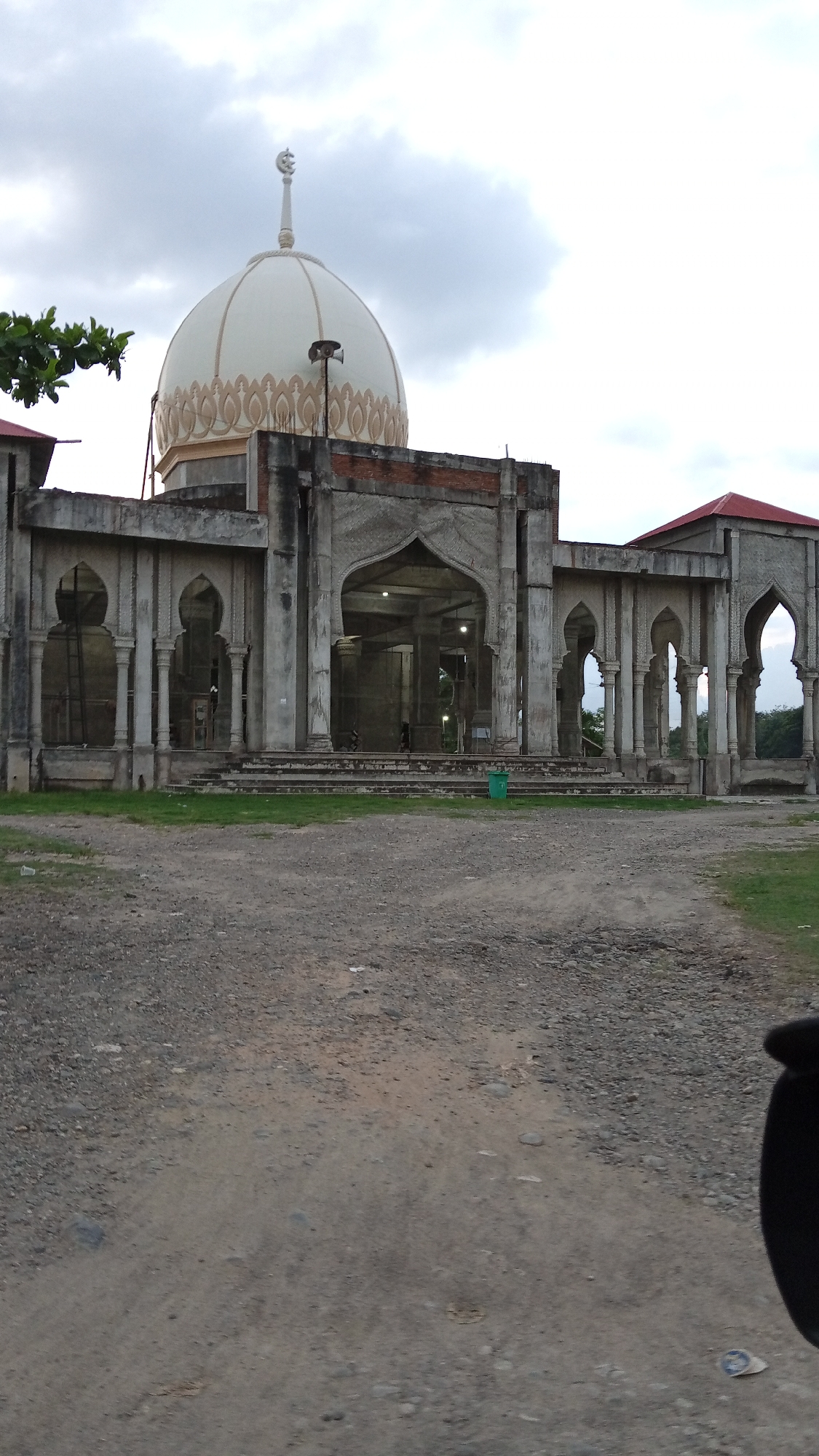 The image shows the partially constructed or dilapidated state of a mosque in Kilometer Delapan, Indonesia. The mosque features a white dome adorned with a pattern. The structure is concrete with arches and pillars, appearing unfinished or possibly abandoned. The foreground consists of a gravelly area, and the sky is overcast. A green trash can stands near the base of the mosque. The atmosphere seems quiet and still, potentially suggesting the site is not currently in use.