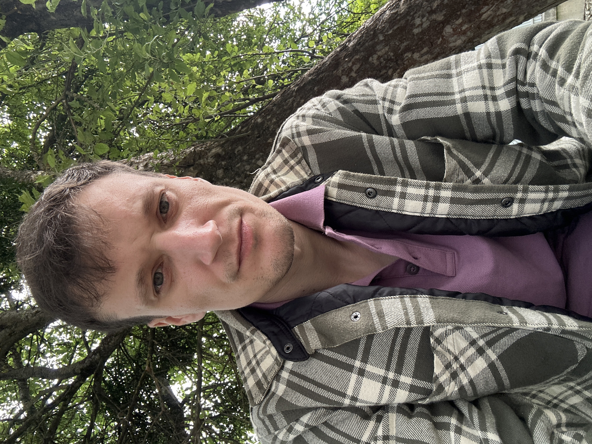 A man is pictured in a selfie taken outdoors, likely in Makiv, Ukraine, leaning against a tree with lush green foliage. He is wearing a plaid jacket over a purple polo shirt. The man has short, brown hair and light-colored eyes. His expression is neutral. The background consists of a large tree trunk and branches with leaves, suggesting a natural, possibly wooded, setting. The lighting indicates it is daytime, with dappled sunlight filtering through the leaves. No other individuals are visible in the image.