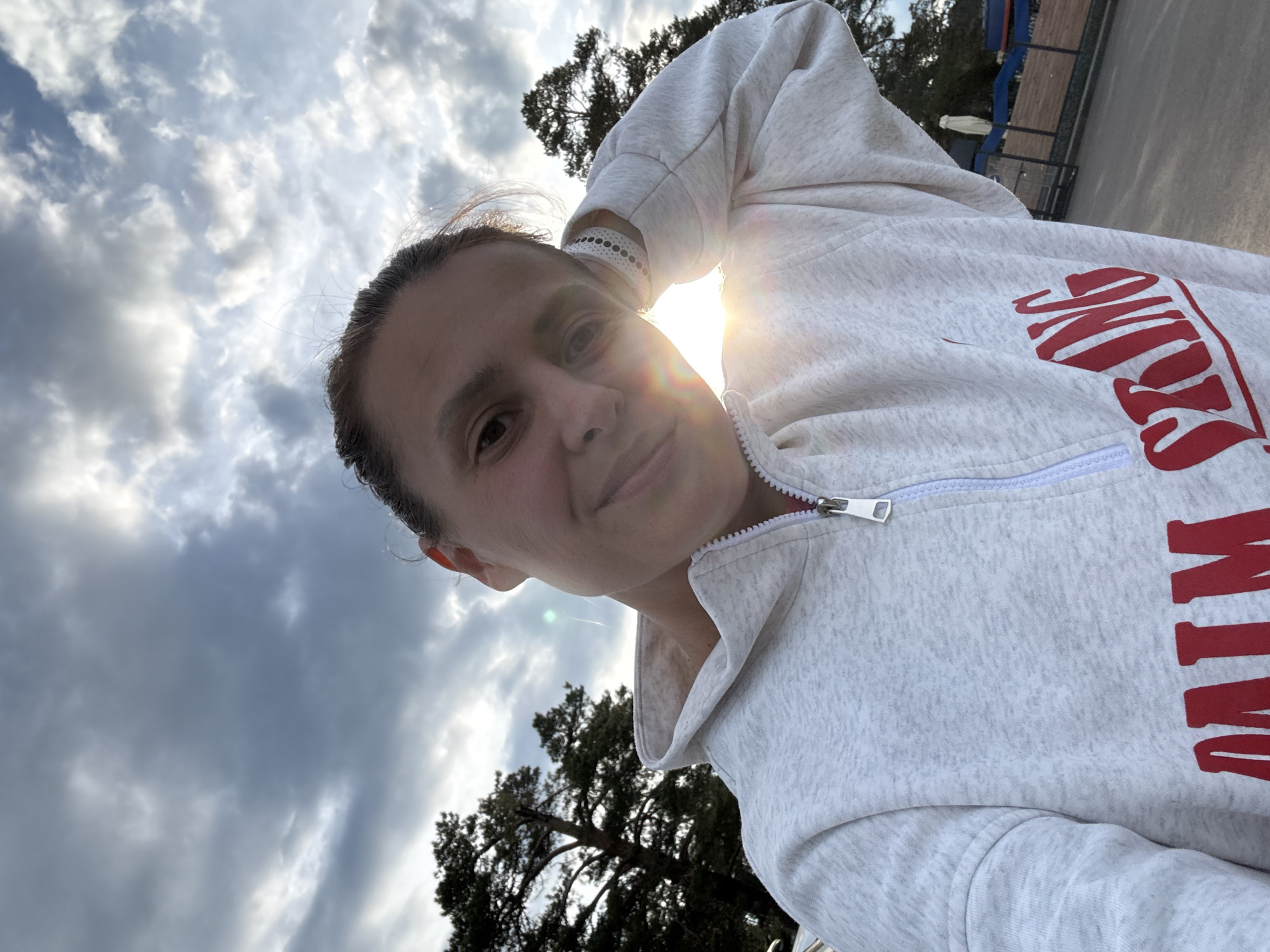 A woman is taking a selfie. She is wearing a gray zip-up hoodie with red lettering and a white patterned wristband. The sun is shining from behind her arm. Behind her, there are trees and a cloudy sky. In the background, there is a structure with blue and white elements and fencing, suggesting a park or recreational area. The location may be Votkinsk, Russia, based on the presence of similar environmental elements found in that region.