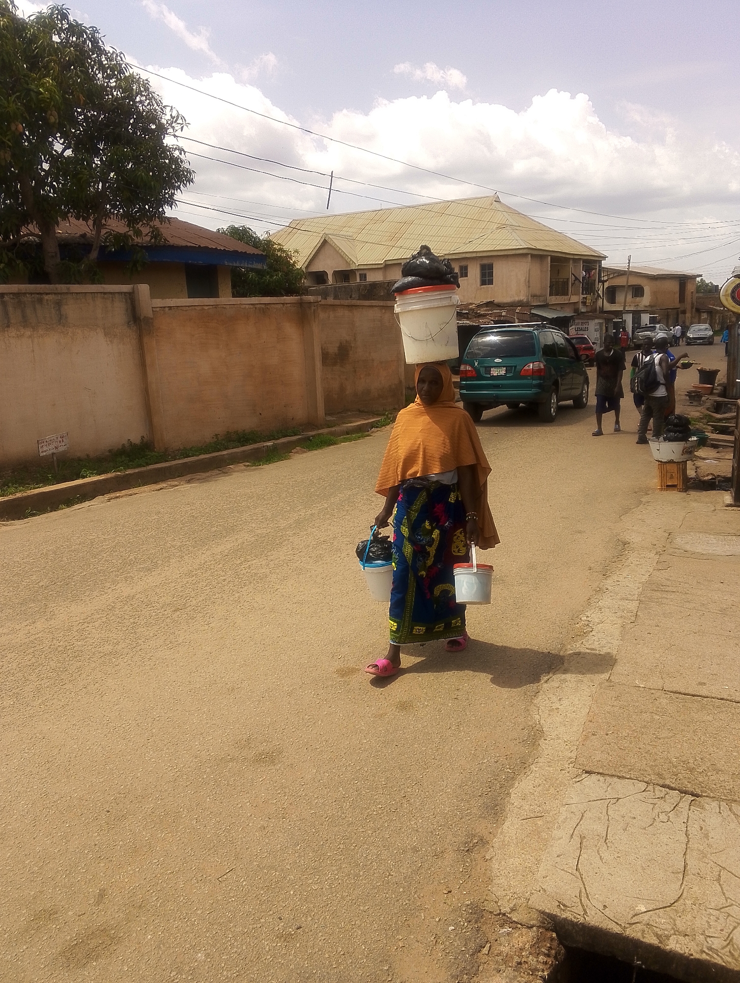 A woman in Jos, Nigeria walks down a paved street carrying three white buckets. One bucket is on her head with black contents inside, and one bucket is carried in each hand. She is wearing pink shoes, a blue and yellow patterned skirt, and an orange head covering and shawl. A tan wall runs along the left side of the road. A green SUV is parked on the right side of the road along with several people standing near a table with a bucket on it. Buildings are visible in the background. The sky is cloudy.