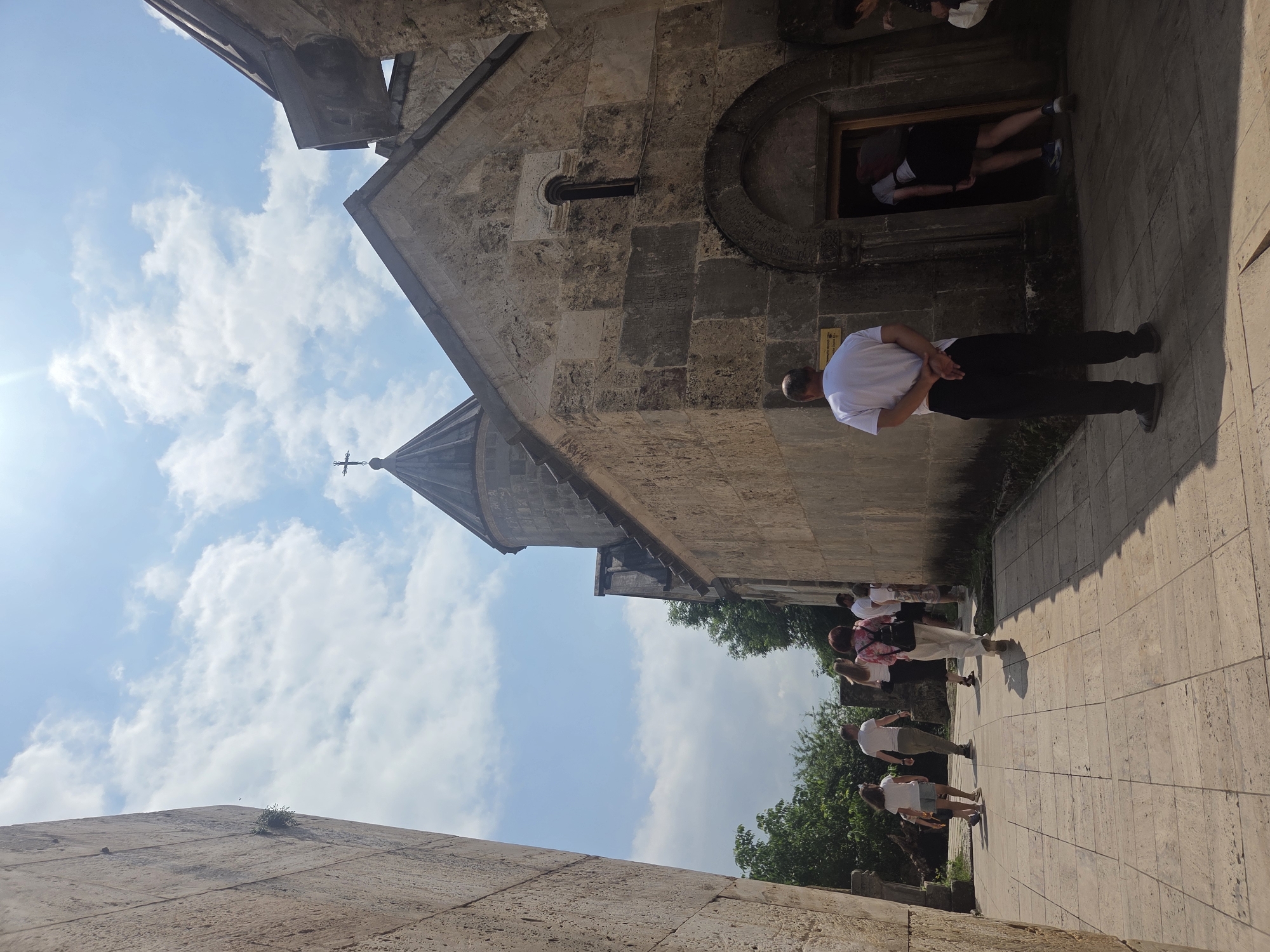 In Teghut, Armenia, a group of tourists walks along a paved path towards a stone church under a bright, cloudy sky. The church, constructed from large, rough-hewn stones, features a conical dome topped with a cross. An arched doorway leads into the building, where a person is visible inside. A man in a white t-shirt and black pants stands facing away from the camera, his hands clasped behind his back, looking towards the church. The path is lined on the left by a tall, textured stone wall. Lush green trees are visible in the background to the left of the church. The sun casts shadows on the ground.