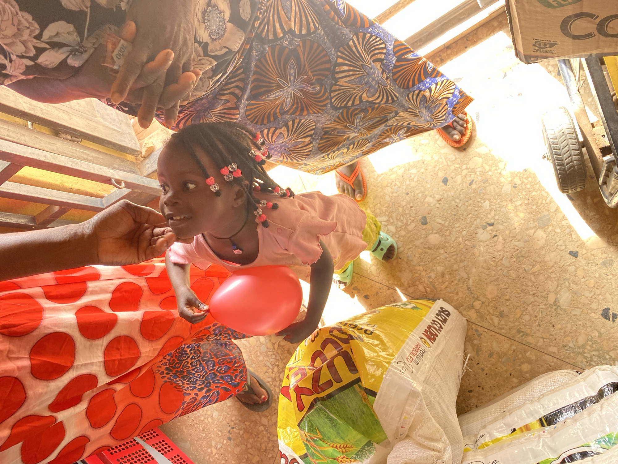 This image captures a vibrant scene in Bukuru, Nigeria, focusing on a young girl interacting with adults in what appears to be a busy, sunlit environment, possibly a market or a shop. The perspective is from slightly above, looking down into the scene.
The central subject is a young girl with dark, braided hair adorned with numerous pink and white heart-shaped beads. She wears a light pink long-sleeved top and looks upwards and to her right with a gentle smile, her eyes wide and engaged. In her left hand, she holds a red, deflated balloon.
Parts of several adults are visible around her. In the upper left, a pair of hands, likely an adult's, holds a folded 1000 Naira banknote. Another adult's hand is seen in the lower left, gently touching the child's chin or face. Colorful patterned fabrics, characteristic of West African attire, are prominent; one garment features blue, orange, and black floral-like patterns, while another in the lower left displays large red polka dots on a white background. An adult's foot wearing an orange flip-flop is also visible on the light-colored, speckled floor.
The setting is brightly lit by strong sunlight, casting defined shadows on the floor. Several bags of goods are scattered on the ground. A prominent yellow and white bag is clearly labeled "SLTY GOLDEN Premium PARBOILED Rice." Another white sack is partially visible. To the right, a cardboard box shows parts of the text "CO" and "ENDORSED BY". The background suggests an open-air or partially covered structure with metal bars.
The overall impression is one of a warm, social interaction, possibly a moment of exchange or gifting, highlighted by the child's joyful expression and the balloon.