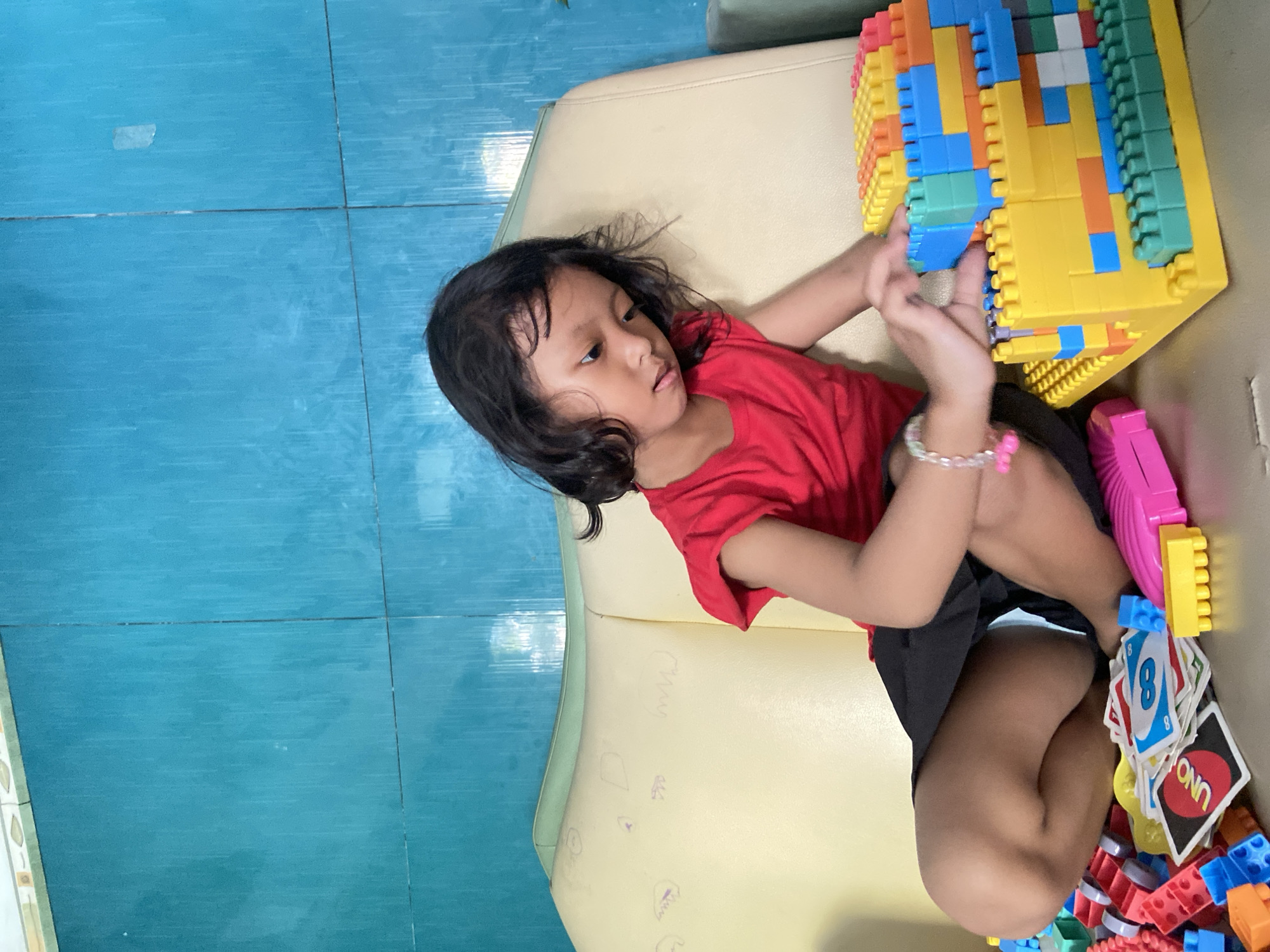 A young girl is lying on a light yellow sofa. She is wearing a red shirt and black shorts. Her hair is dark and slightly messy. She is playing with colorful plastic building blocks, which appear to be stacked into a rectangular tower. There are also Uno cards and more building blocks scattered near her legs. The floor is tiled with blue tiles. There is no indication from the image that the subject is in the city of Gelam, Indonesia.
