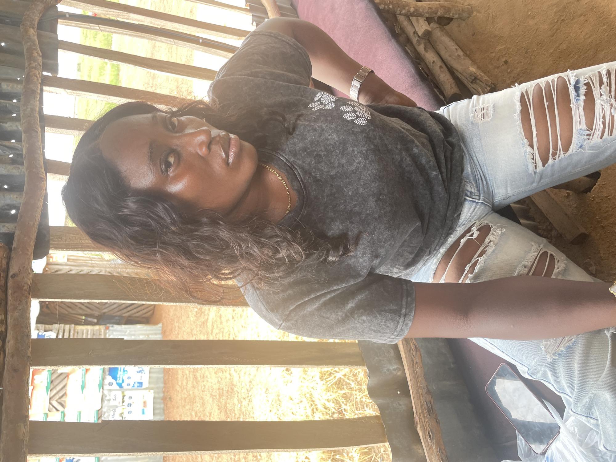 A young woman with dark, wavy hair is posing for a photo in a rustic, open-air structure, likely a shed or makeshift shelter. She is wearing a dark grey, acid-wash t-shirt with a subtle white graphic on the chest and light blue, ripped jeans. Her arms are adorned with a silver watch on her left wrist and a thin gold chain around her neck. She is reclining, with her head propped up, and looking directly at the camera with a serene expression.
The setting appears to be outdoors, with natural light streaming in, suggesting it is daytime. The structure is made of unfinished wood and corrugated metal. Behind the woman, there are visible wooden slats and some objects that might be sacks or bundles. The ground is dirt. In the background, through the wooden slats, glimpses of green vegetation and more rustic structures can be seen. A pink mat or cushion is visible beneath her. A mobile phone, with a pink casing, lies near her right leg. The overall impression is one of a casual, candid moment captured in a rural or undeveloped area.