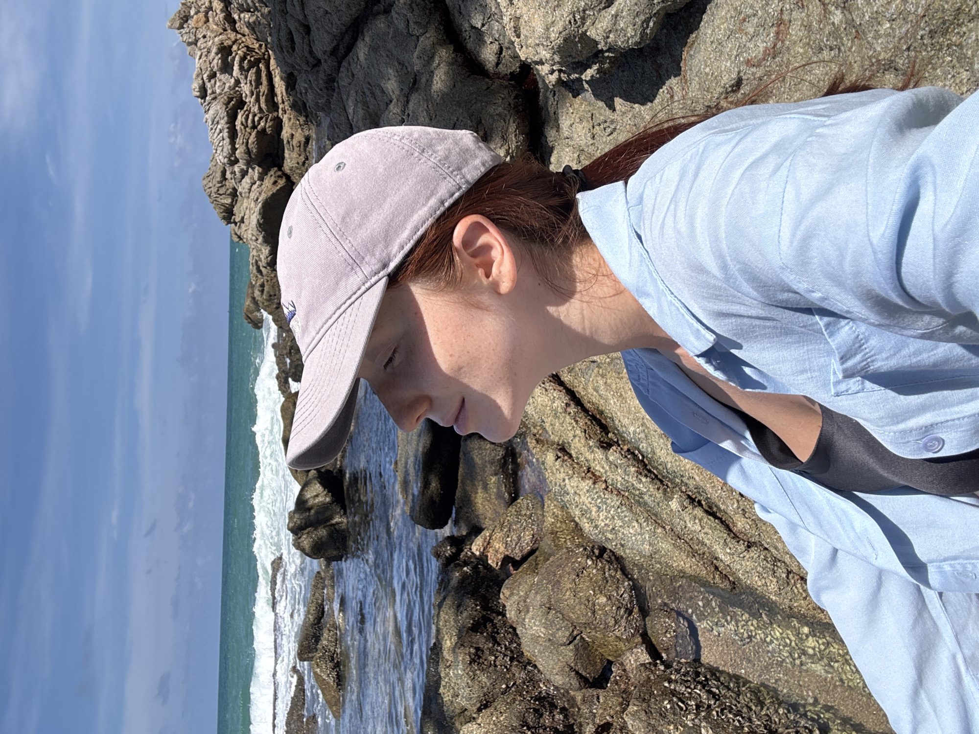 The image depicts a person wearing a light blue collared shirt and a light purple baseball cap. The individual is positioned amongst rocks near a body of water with waves. The location appears to be a coastal area, based on the presence of the ocean. The image was taken in Choeng Thale, Thailand.