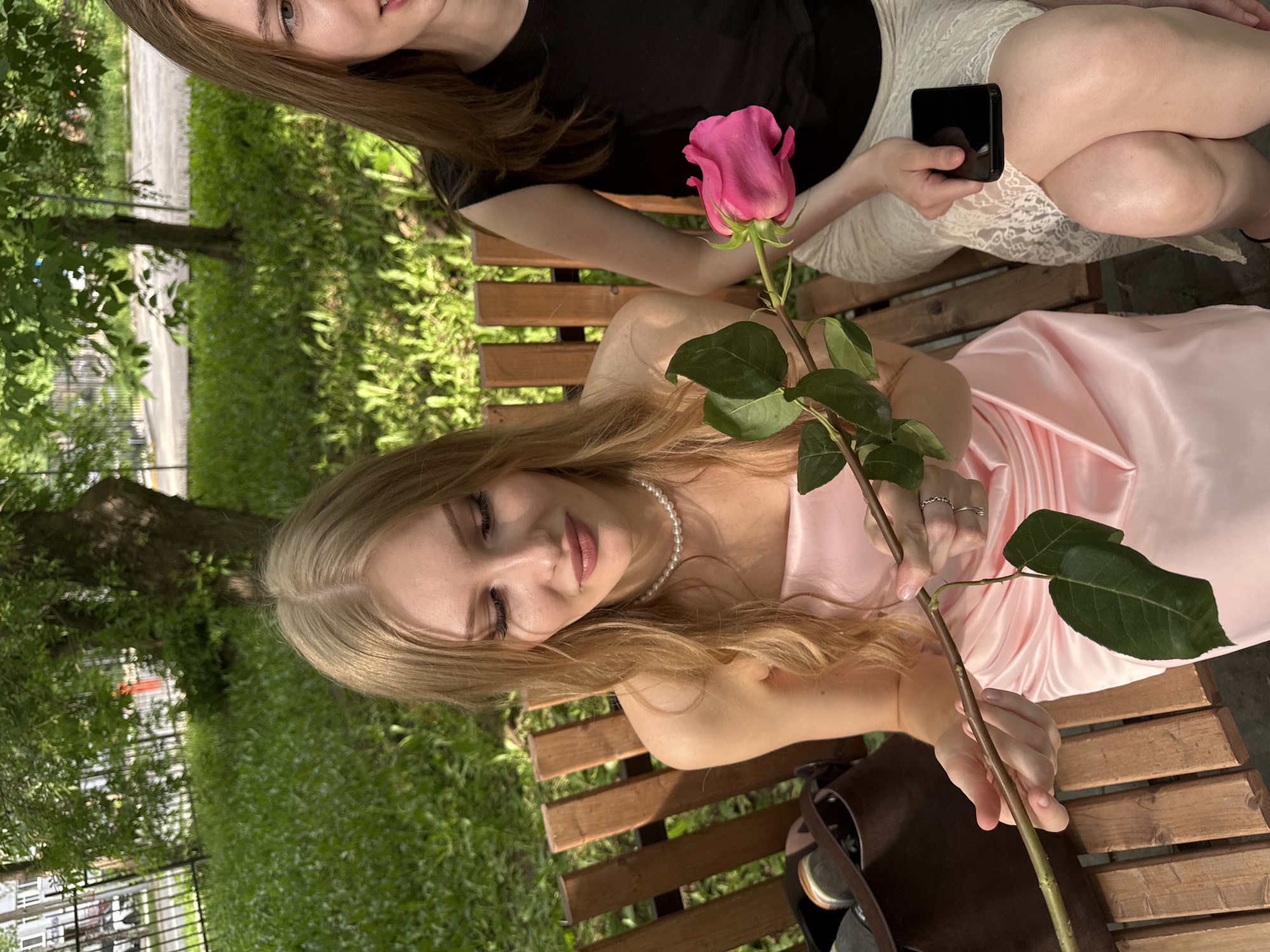 A medium, high-angle shot reveals two women sitting on a wooden park bench in a green, outdoor setting. The woman in the foreground, dressed in a pink satin dress, holds a long-stemmed pink rose, gazing down at it. She wears a pearl necklace and several rings. A dark brown bag sits beside her on the bench. Behind her, another woman in a black shirt and white lace skirt sits with a smartphone in her hand. Green grass and trees surround the bench. Based on the visual information, the location can't be accurately identified as Perm, Russia, but it appears to be a park or garden setting.