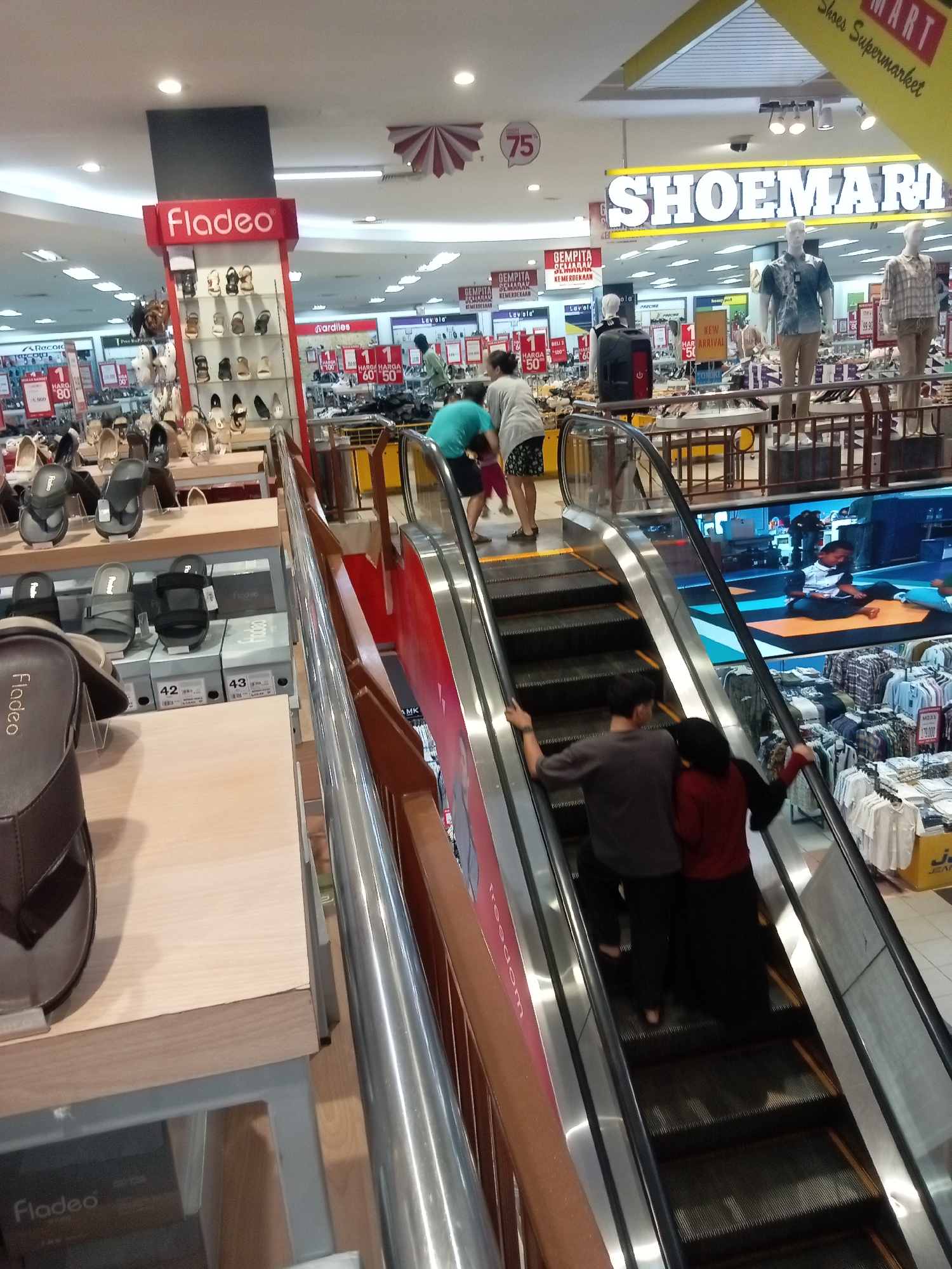 The image shows an interior view of a Shoemart department store, likely located in Batam, Indonesia, as indicated by the signage and store layout typical of Indonesian retail environments. The scene captures a section of the store featuring an escalator and surrounding merchandise displays.
In the foreground, on the left, is a display of brown men's sandals with the brand name "Fladeo" visible on a price tag. Above this, more shoes are displayed on shelves, including sandals and heels, also branded "Fladeo".
The main focus of the image is the escalator in the center, moving upwards. Two people are ascending the escalator: a man wearing a dark gray t-shirt and black pants, holding the handrail, and a woman behind him in a red long-sleeved shirt and black skirt, also holding the handrail. Their interaction suggests they are together. Further up the escalator, another person in a turquoise shirt is visible.
The background reveals the broader store environment. To the left of the escalator, a large "Fladeo" sign is prominent, with more shoe displays. Across from the escalator, a large sign reads "Shoemart". Further into the store, mannequins are displayed wearing casual shirts and pants, and various racks of clothing and shoes are visible. Signs advertising promotions, such as "HARGA 80" and "NEW ARRIVAL", are interspersed throughout the displays. The lighting is typical of an indoor retail space, with overhead fluorescent lights illuminating the scene. The floor appears to be tiled. The overall impression is one of a busy retail environment.