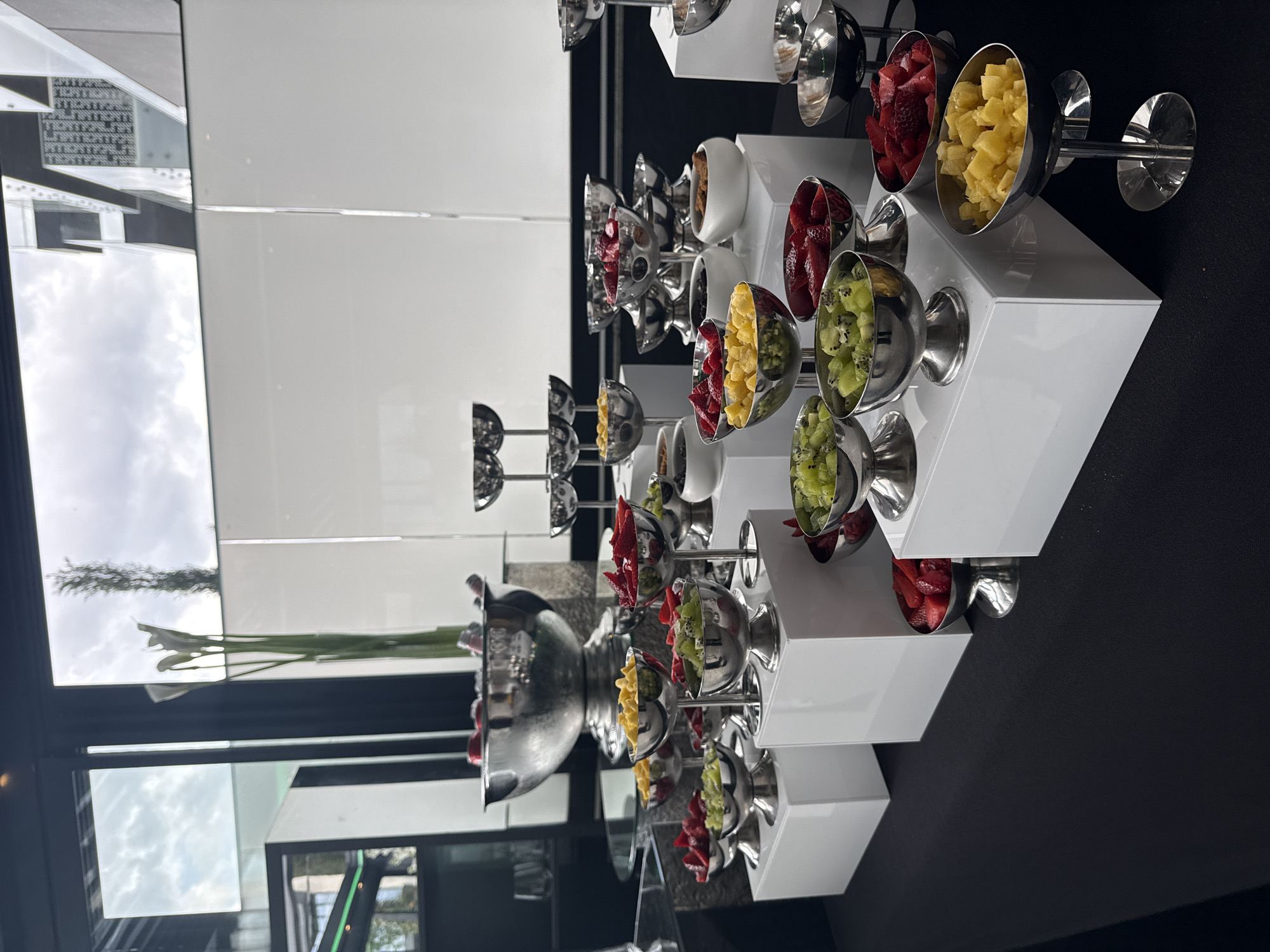 The image displays a modern and elegant fruit bar or buffet station, likely set up indoors at a restaurant, hotel, or event venue. The main subjects are various types of pre-cut fresh fruit presented in numerous polished silver bowls. These bowls are arranged on sleek, tiered white display stands, creating an attractive and organized presentation.
Visible fruits include diced strawberries (red), cubed pineapple (yellow), and chopped kiwi (green). Some white ceramic bowls are also visible, possibly containing other toppings like nuts or granola, though their contents are not clearly identifiable. A large silver bowl or vase on the left side of the display holds tall green stalks or foliage, adding a decorative element.
The activity taking place is the display of a selection of fruits for serving, suggesting a self-service buffet or dessert station. The setting appears bright, indicating it's daytime, with ample natural light. Large reflective surfaces, possibly mirrors or windows, are prominent in the background, showing clear reflections of a cloudy sky and some distant structures, implying an outdoor view. The display rests on a dark, possibly black, surface.
Notable details include the uniformity of the silver bowls and white stands, contributing to a clean and sophisticated aesthetic. The fruits appear fresh and vibrantly colored. There is some text visible in reflections on the left side of the image, but it is too small and distorted to be legible. There are no specific visual cues to confirm the location as Sofia, Bulgaria, beyond the general context provided.