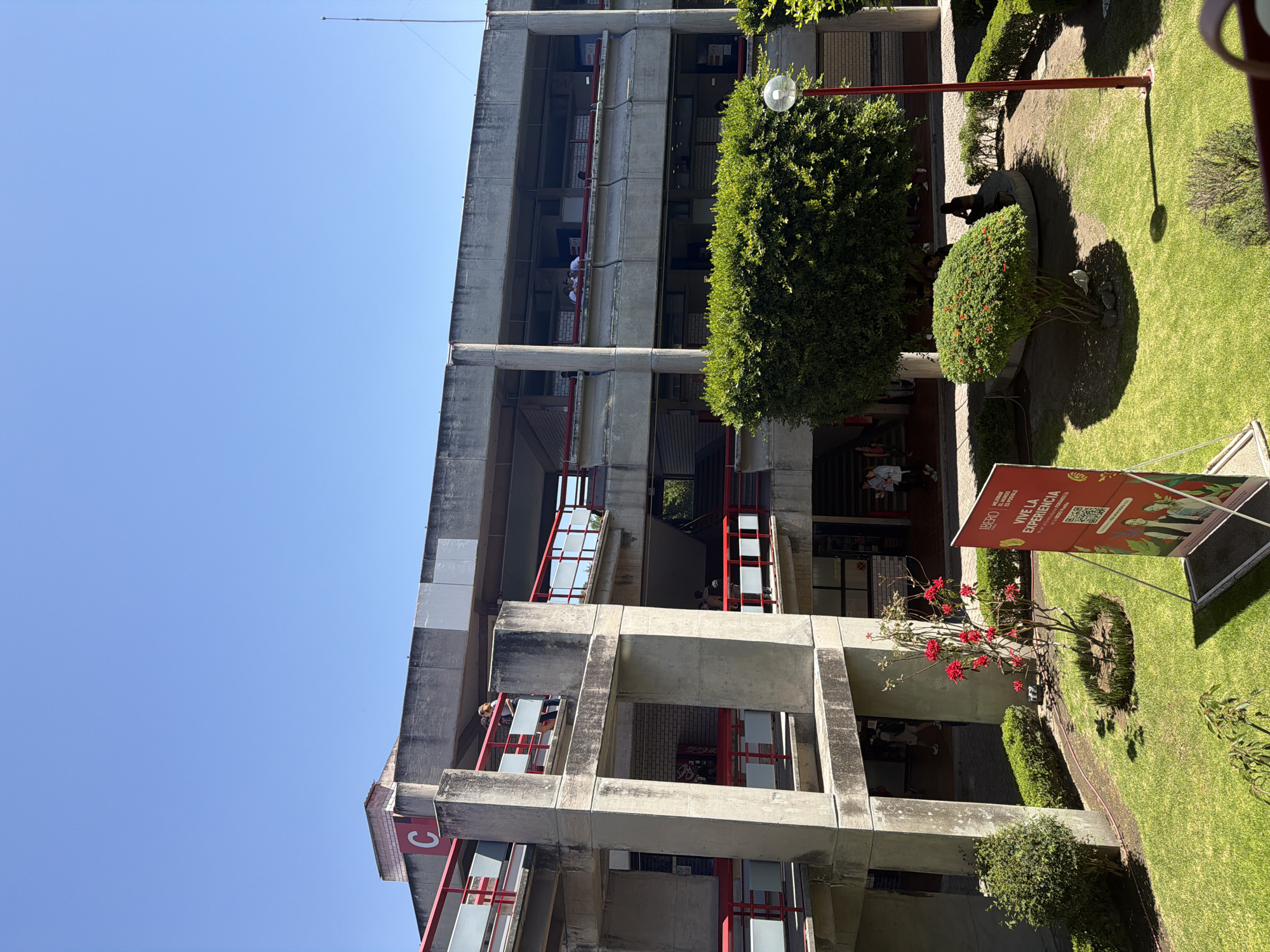 The image captures a bright, sunny outdoor scene at a university campus, likely in San Bernardino Tlaxcalancingo, Mexico. A prominent multi-story concrete building, exhibiting a modernist or brutalist architectural style with open-air corridors and red railings, occupies the left side. Numerous people are visible on different levels of the building, either standing or seated, engaging in typical campus activities. A large white letter "C" on a red background is visible on one section of the building.
The right side of the frame features a well-maintained green lawn interspersed with various plants and trees. A tall, conical evergreen tree and several manicured bushes are present, alongside a plant with vibrant red flowers. A dark, abstract statue or sculpture is situated within a circular patch in the grassy area. A street lamp with a spherical light source stands near the evergreen tree. Strong shadows cast across the lawn and structures indicate bright daylight.
A red banner stands in the foreground on the right, featuring the "IBERO" logo and the Spanish text "VIVE LA EXPERIENCIA DE LA UNIVERSIDAD QUE QUIERES" (Live the experience of the university you want), along with a QR code and illustrations, advertising Universidad Iberoamericana. The sky is clear and bright blue, and an antenna extends from the building's roof towards the top of the frame. The overall atmosphere appears calm and active, characteristic of a university setting during a clear day.