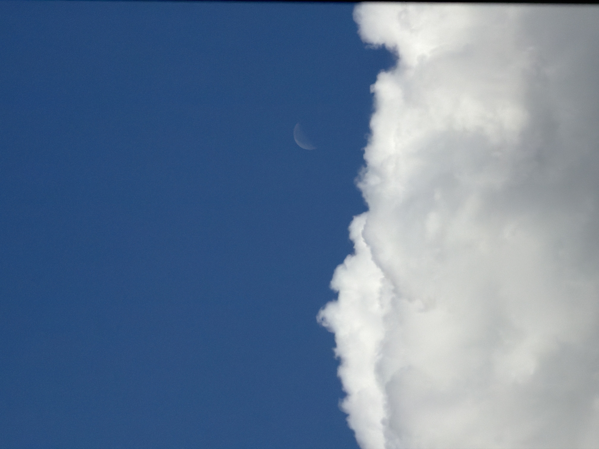 A daytime shot captures a blue sky and white clouds. A crescent moon is visible in the blue sky. The clouds occupy the right side of the frame, creating a sharp contrast with the clear blue sky to the left.
While I can describe the scene, I cannot confirm its location in Sofia, Bulgaria, as there are no geographical markers in the image.