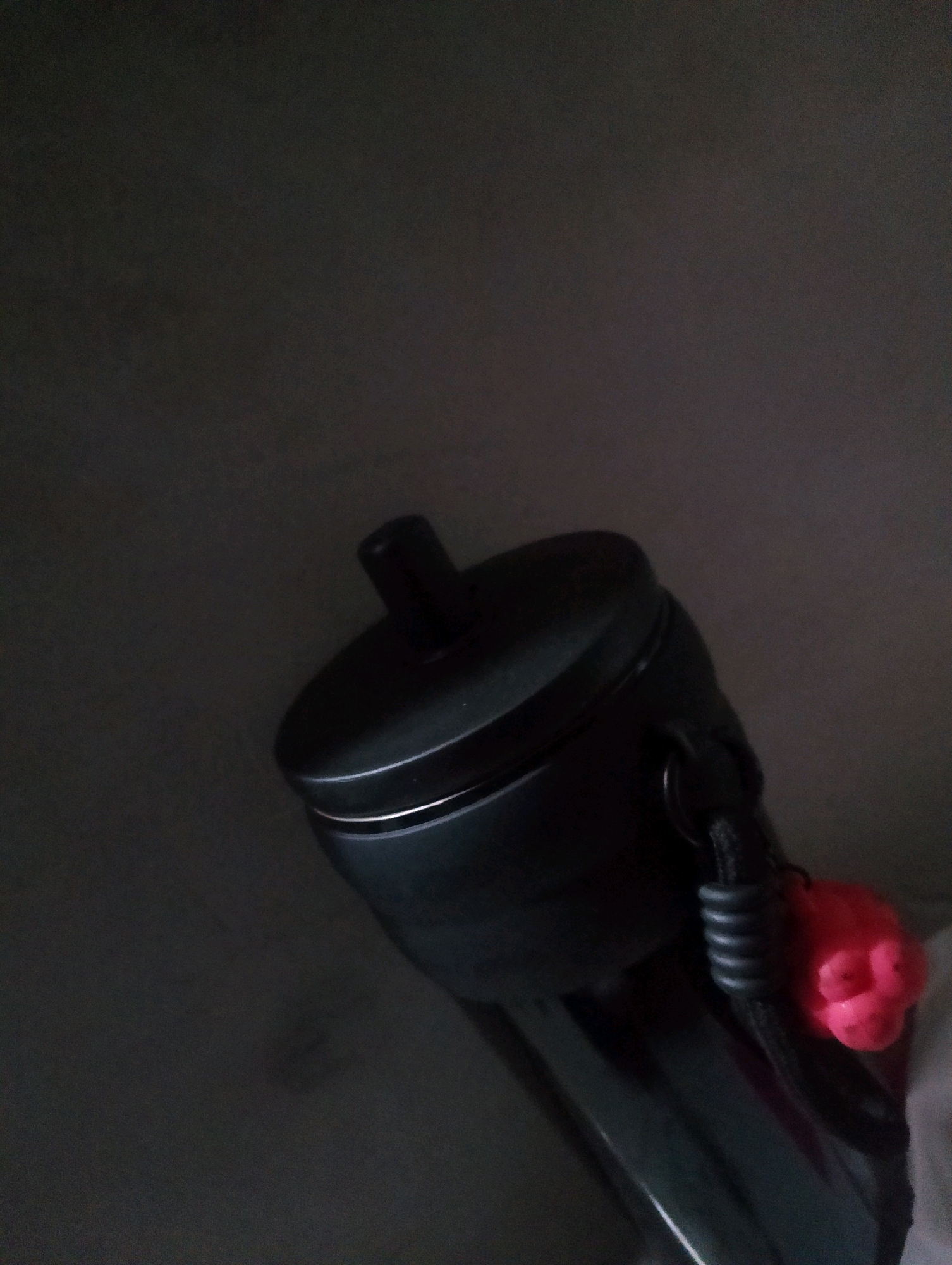 A black flashlight is positioned diagonally in the frame, with its top portion slightly out of focus. Attached to the flashlight is a coiled black strap, which leads to a small, bright pink, face-like charm. The background is dark and indistinct, with subtle lighting suggesting an indoor environment. The location is not specified.