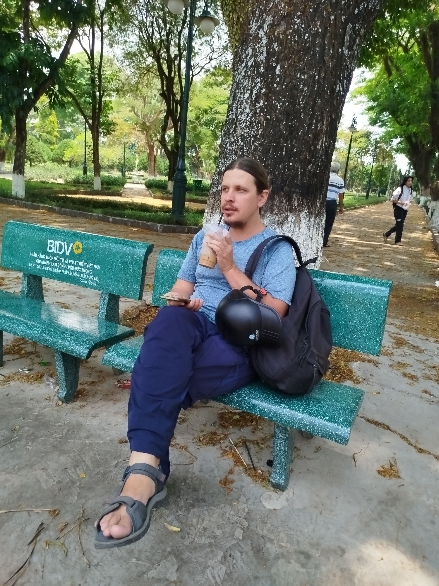 A man is sitting on a green park bench holding a drink and a mobile phone. He has a backpack and a helmet resting next to him on the bench. The bench has the logo and name of BIDV, a Vietnamese investment and development bank, as well as text referencing the branch of Lâm Đồng in Đức Trọng. The text further specifies that the location is at the Liên Nghĩa Market intersection of Phạm Văn Đồng and Trần Hưng Đạo streets in Liên Nghĩa, Vietnam. Behind the man is a large tree and other park trees. Two people are walking on a path in the background.