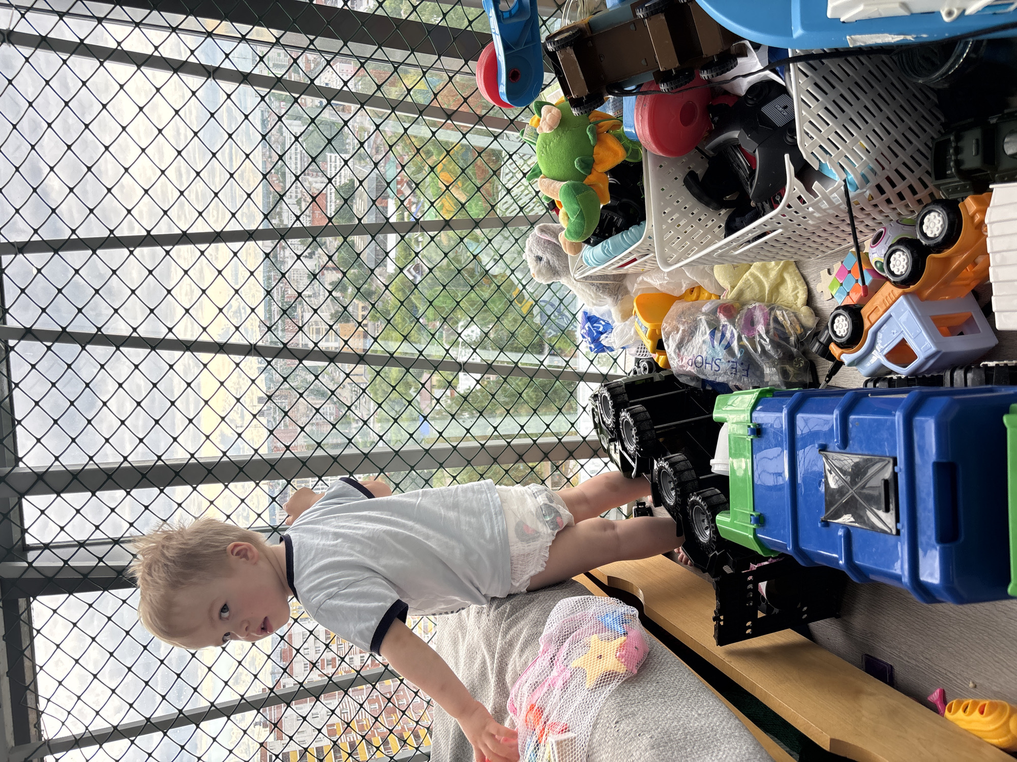 A child lies on their back on a gray upholstered surface, facing the camera. The child wears a light blue t-shirt with dark blue trim and a diaper. Their bare feet are up in the air, near the wheels of a large blue and green toy truck. Behind the child, a wire safety barrier obscures the view of a city landscape with buildings, trees, and cloudy sky. A variety of toys are piled behind the child and the truck. The location is not Izhevsk, Russia.
