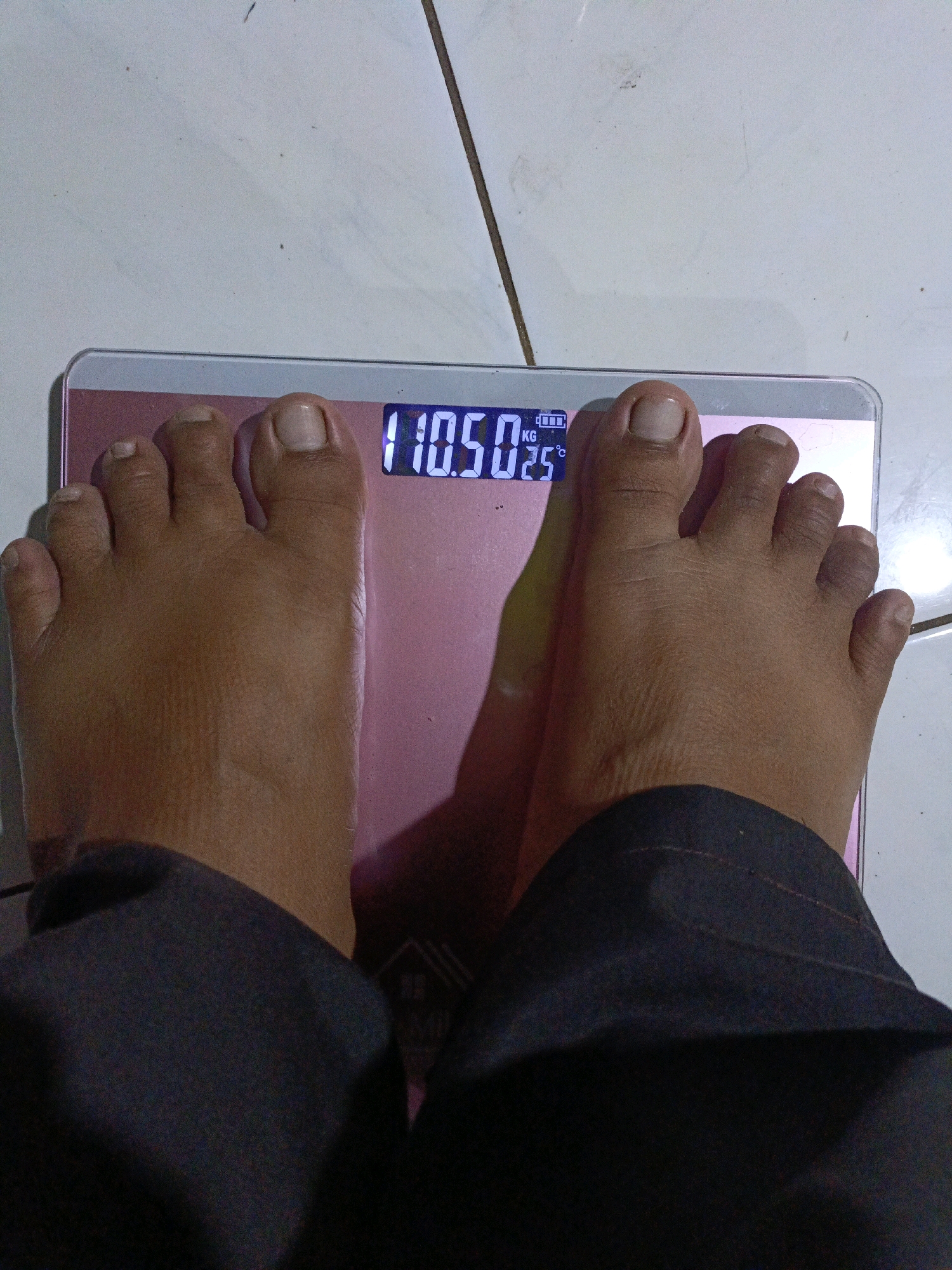 An overhead, medium shot captures a person's feet standing on a pink digital scale, which displays a weight of 110.5 kg and a temperature of 25°C. The scale rests on a tiled floor. The person is wearing dark pants that obscure their legs from the shins down. The lighting is artificial, casting shadows from the feet onto the scale. The scene is likely a private residence, given the domestic setting of a scale on a tiled floor. There are no discernible landmarks to pinpoint a specific location within Jenu, Indonesia.