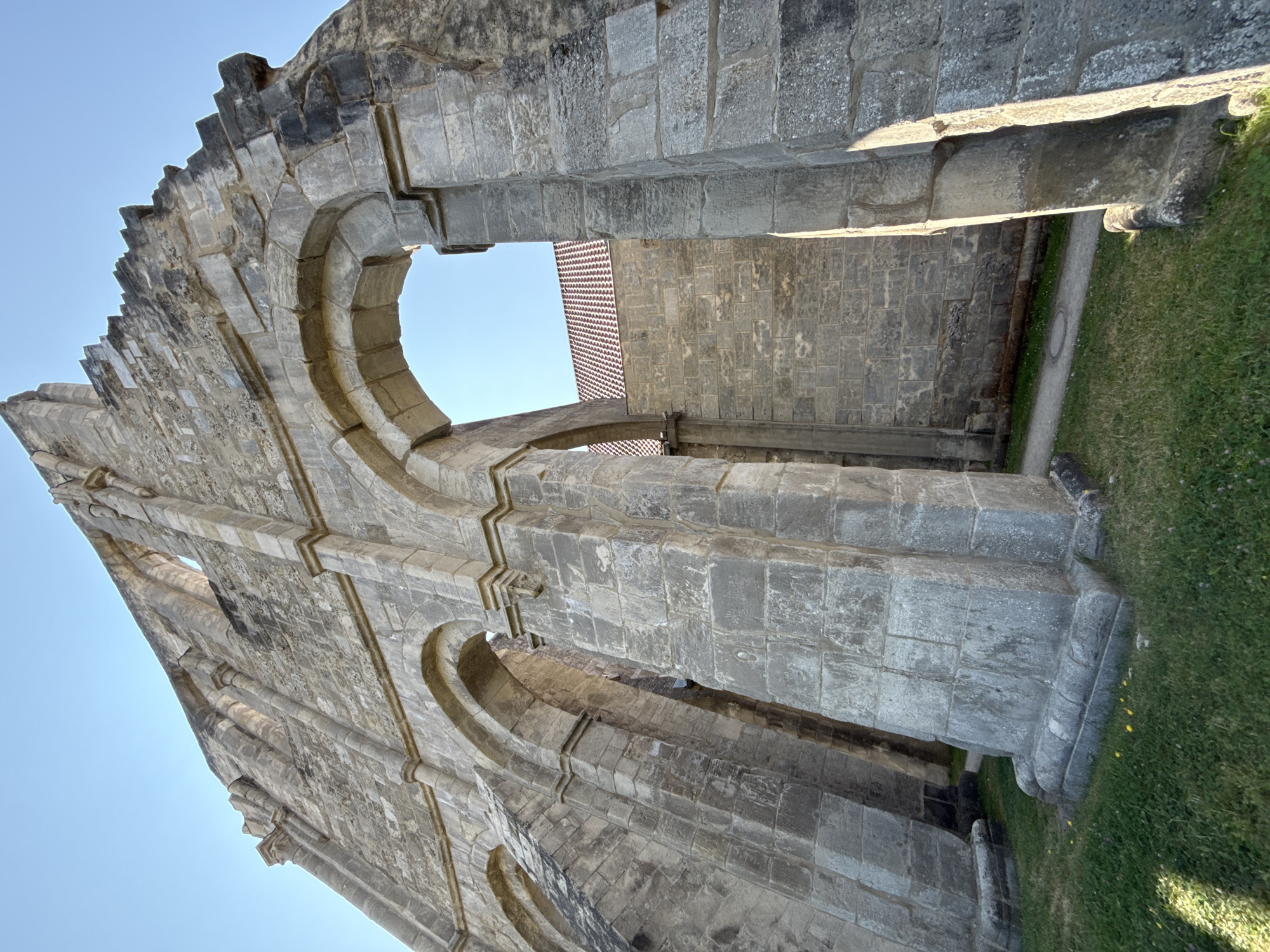 The image shows the ruins of the Walkenried Abbey in Walkenried, Germany, under a clear blue sky. The perspective is low, looking up at the massive stone arches and walls of the ancient structure. The stone is weathered and appears to be a light gray, with some darker patches. The arches are rounded and supported by thick, segmented pillars. Beyond the arches, another stone wall with a tiled roof is visible. The ground in front of the ruins is covered in green grass, with a few small yellow wildflowers scattered about. A pathway is visible behind the arches. There are no people or animals in the image. The lighting suggests it is either morning or late afternoon, with sunlight casting shadows on the stone.