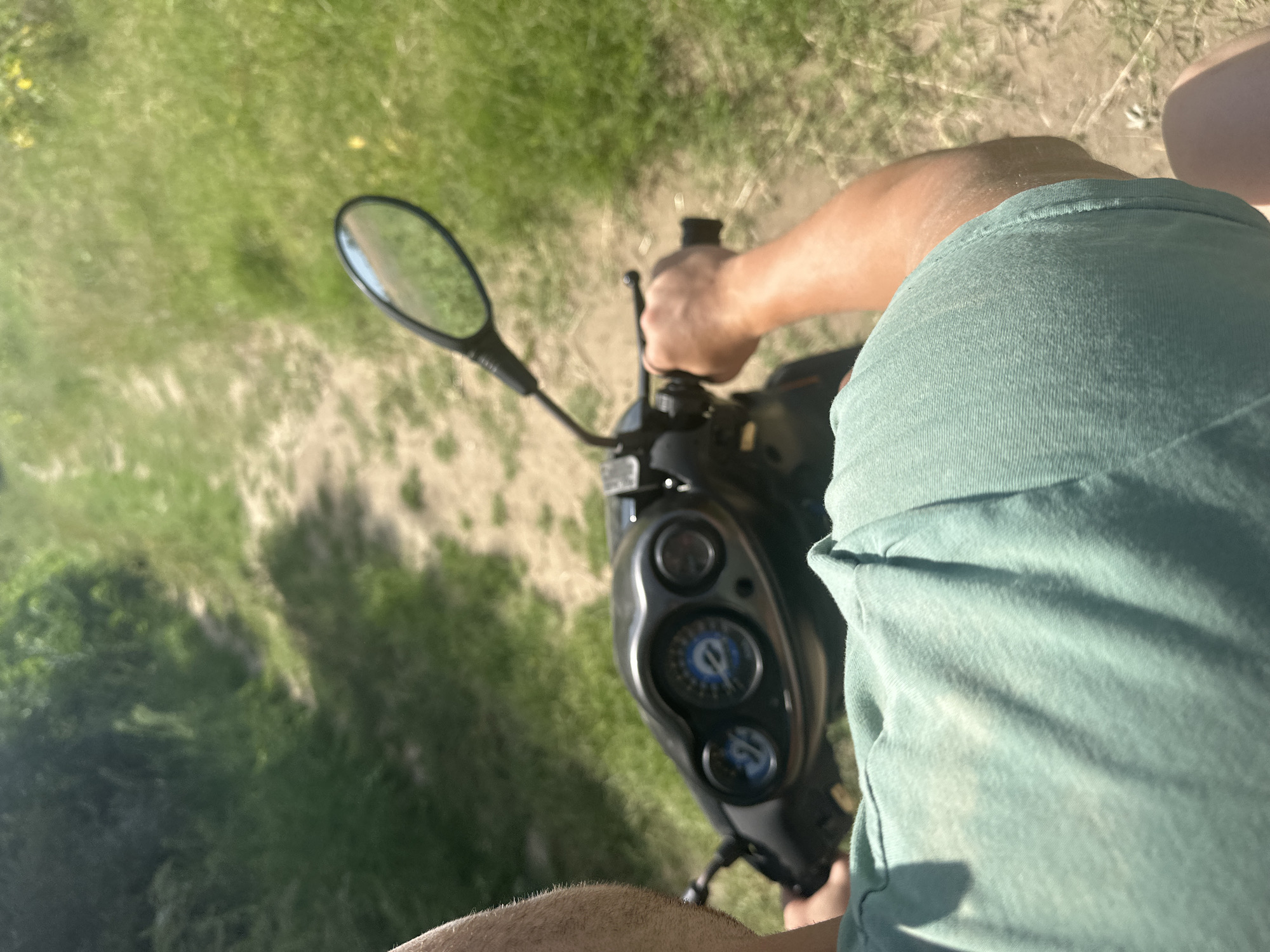A first-person perspective shot shows a person riding a scooter on a dirt path in a grassy area. The rider is wearing a light green t-shirt. The scooter's handlebars and dashboard are visible in the foreground, with a rearview mirror reflecting the path ahead. The path is narrow and winds through tall green grass and some trees in the background. The sun is shining, casting shadows on the path. The location is not identifiable as Malevo, Bulgaria from the image.
