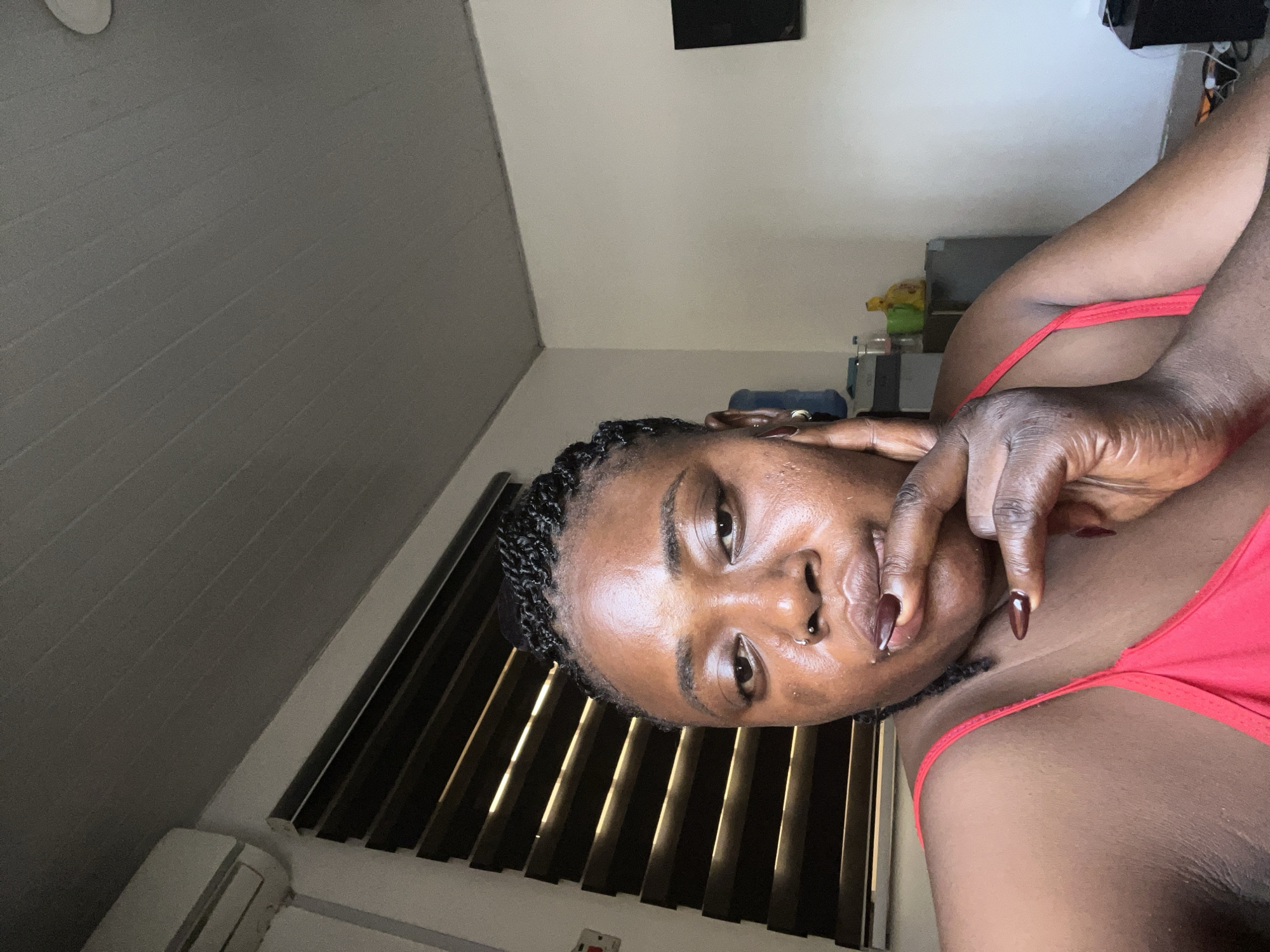 An indoor, eye-level, medium-shot photograph shows a Black woman with braided hair and a nose ring posing in what appears to be a residential setting in Abuja, Nigeria. She is wearing a red spaghetti strap top and her right hand is raised to her mouth, with her index finger on her lips and her thumb resting on her chin. Her fingernails are painted dark brown. She has on a nose ring and small hoop earrings. The background includes a white wall, a dark window blind with horizontal slats, and a white air conditioning unit mounted high on the wall. The ceiling is made of white, slatted panels. To the right of the woman, a portion of a black television screen is visible. A blue water dispenser is visible behind the woman's head.