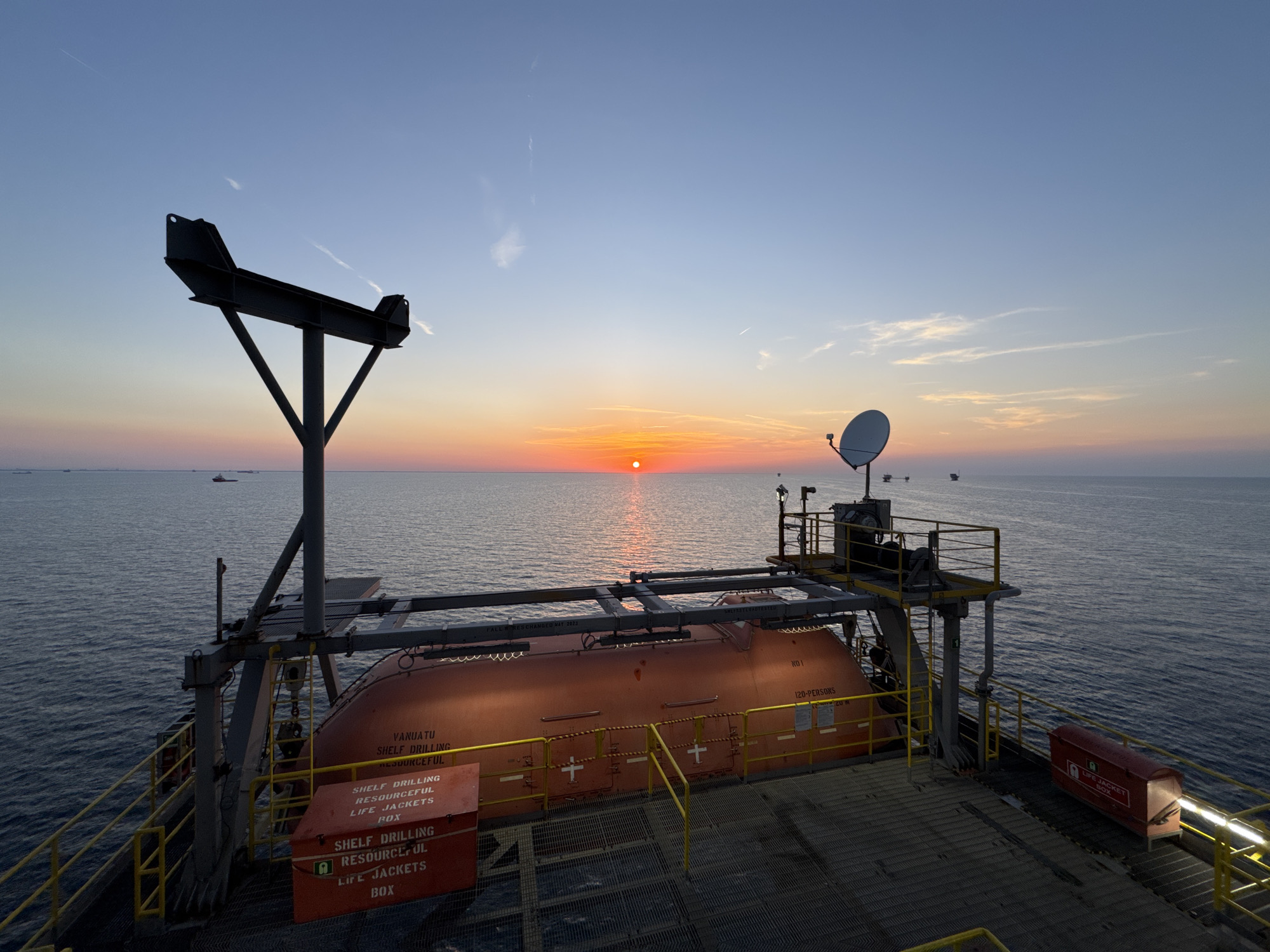 This is a wide-angle, outdoor, horizontal photograph taken at sunset on the ocean. The sun is low on the horizon, casting a warm orange glow across the water and sky. In the foreground, a life raft is suspended from a metal davit system. The life raft is bright orange and appears to be a covered, enclosed type. There are several red boxes labeled "LIFE JACKETS BOX" on the deck. A satellite dish is mounted on a platform to the right of the life raft. The ocean is calm, with small waves. In the distance, several boats and possibly oil rigs can be seen. The sky transitions from a light blue at the top to shades of orange and pink near the horizon. The overall mood of the image is serene and industrial. The location is stated as Unknown, Italy, but the visual evidence points to an offshore industrial setting, not a city.
