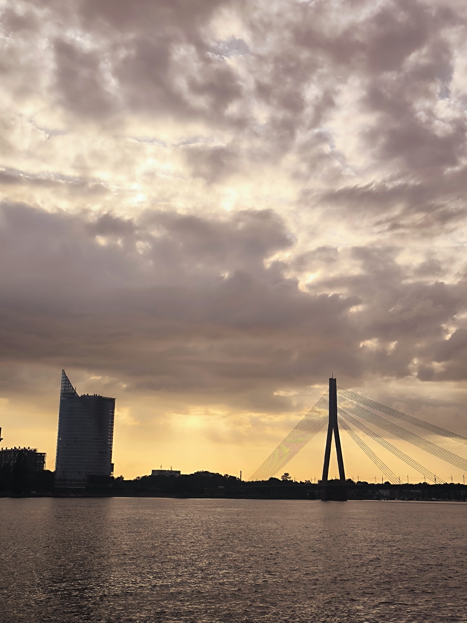 This is a vertical, eye-level, full shot of the Daugava River in Riga, Latvia, at sunset. A large, modern skyscraper, possibly the Parex Bank building, is silhouetted against the hazy, orange-tinted sky on the left. In the center of the frame, the distinctive A-shaped pylon of the Vanšu Bridge, a cable-stayed bridge, dominates the horizon, with its cables fanning out across the sky. The river in the foreground is dark and rippled, reflecting the muted colors of the sky. The sky itself is filled with dramatic, layered clouds, ranging in color from deep purple to soft pink and pale yellow where the sun is setting. The overall impression is one of urban grandeur meeting the natural beauty of the twilight sky. There are no people visible in the image.