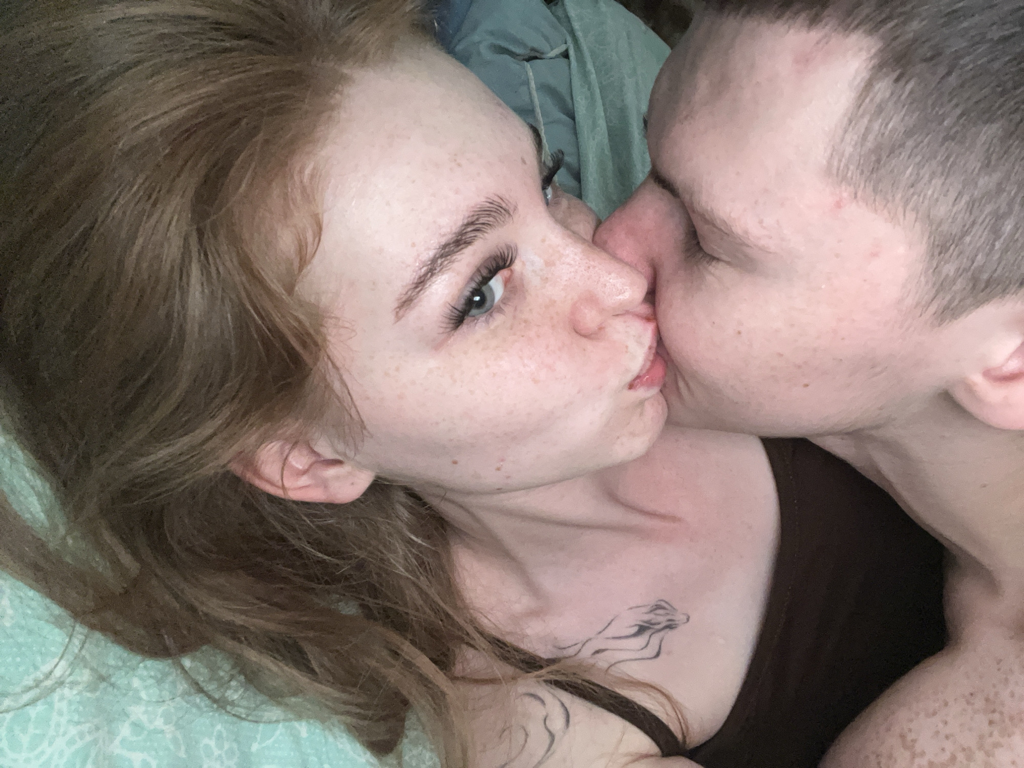 The image is a close-up, overhead shot of a young woman and a young man. The woman, with long, light brown hair and fair skin with freckles, is on the left, facing towards the right. She has on false eyelashes and her left eye is open, looking at the camera. Her lips are slightly parted. The man is on the right, kissing the woman on her cheek. He has short, dark hair and fair skin. He is wearing a dark tank top. A tattoo of a stylized animal is visible on the woman's chest. The background appears to be a bed with a teal and white patterned sheet and a crumpled green fabric. The image does not provide any information or context related to the city of Красные Баки, Russia.
