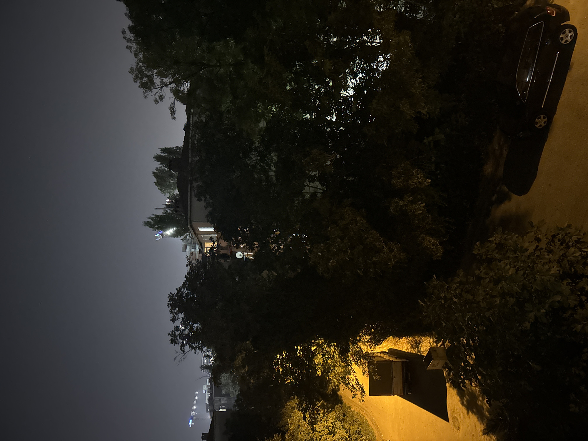 The image depicts an urban nighttime scene in Güngören, Turkey. A street is illuminated by artificial lights, casting long shadows. In the foreground, a dark-colored car is parked on the right side of the street. To the left, a delivery truck is positioned on the road. The background features buildings and trees, with some construction cranes visible in the distance. The sky is dark, indicating it is nighttime. There are no people visible in the image. The overall impression is of a quiet urban environment after dark.