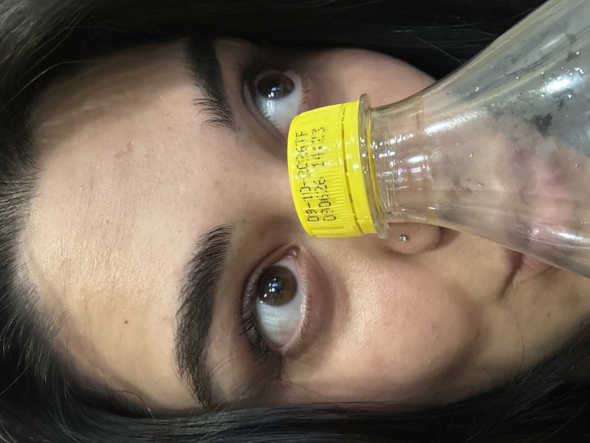 An extreme close-up shot focuses on the face of a person with dark hair, partially obscured by a clear plastic bottle with a yellow cap. The person's right eye, with a brown iris and long eyelashes, is clearly visible and looking to the left of the frame. A small piercing is visible on the person's left cheekbone, near the nose. The yellow bottle cap has black text printed on it, which appears to be a date and time stamp. The background is blurred and dark, suggesting an indoor setting with low lighting. The overall impression is one of candidness and informality.