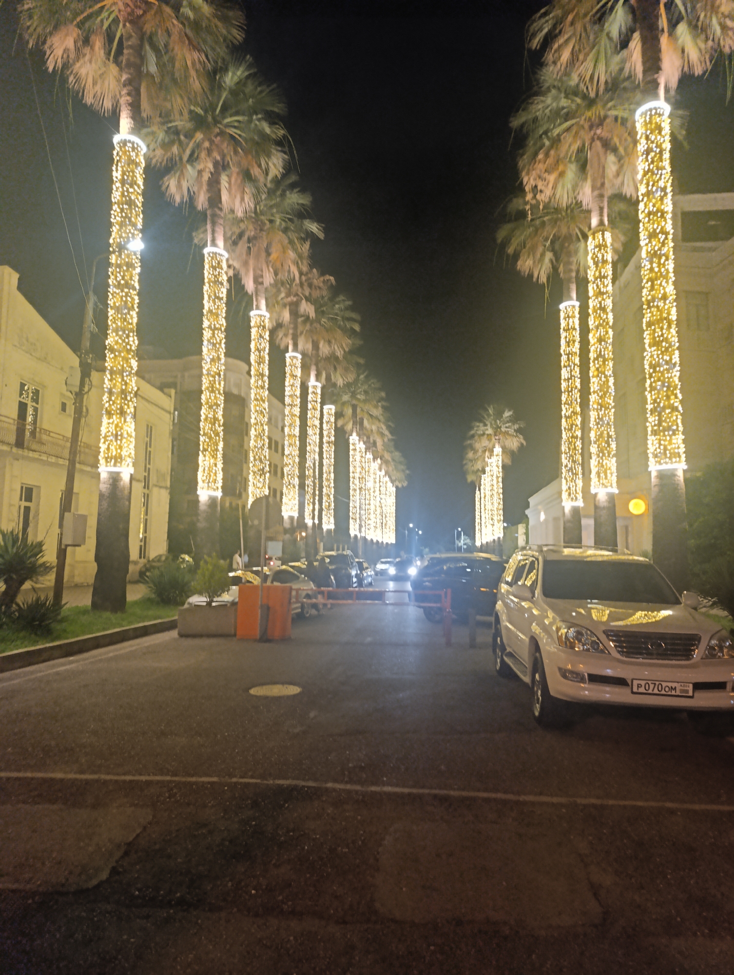 The image shows a street in Sokhumi, Abkhazia, at night, decorated with palm trees wrapped in yellow lights. Several cars are parked along the street, and a white SUV is in the foreground. Buildings line the street, one of which is yellow and appears to be an apartment building or hotel. The street is paved and appears to be wet, possibly from recent rain. The sky is dark, indicating nighttime.