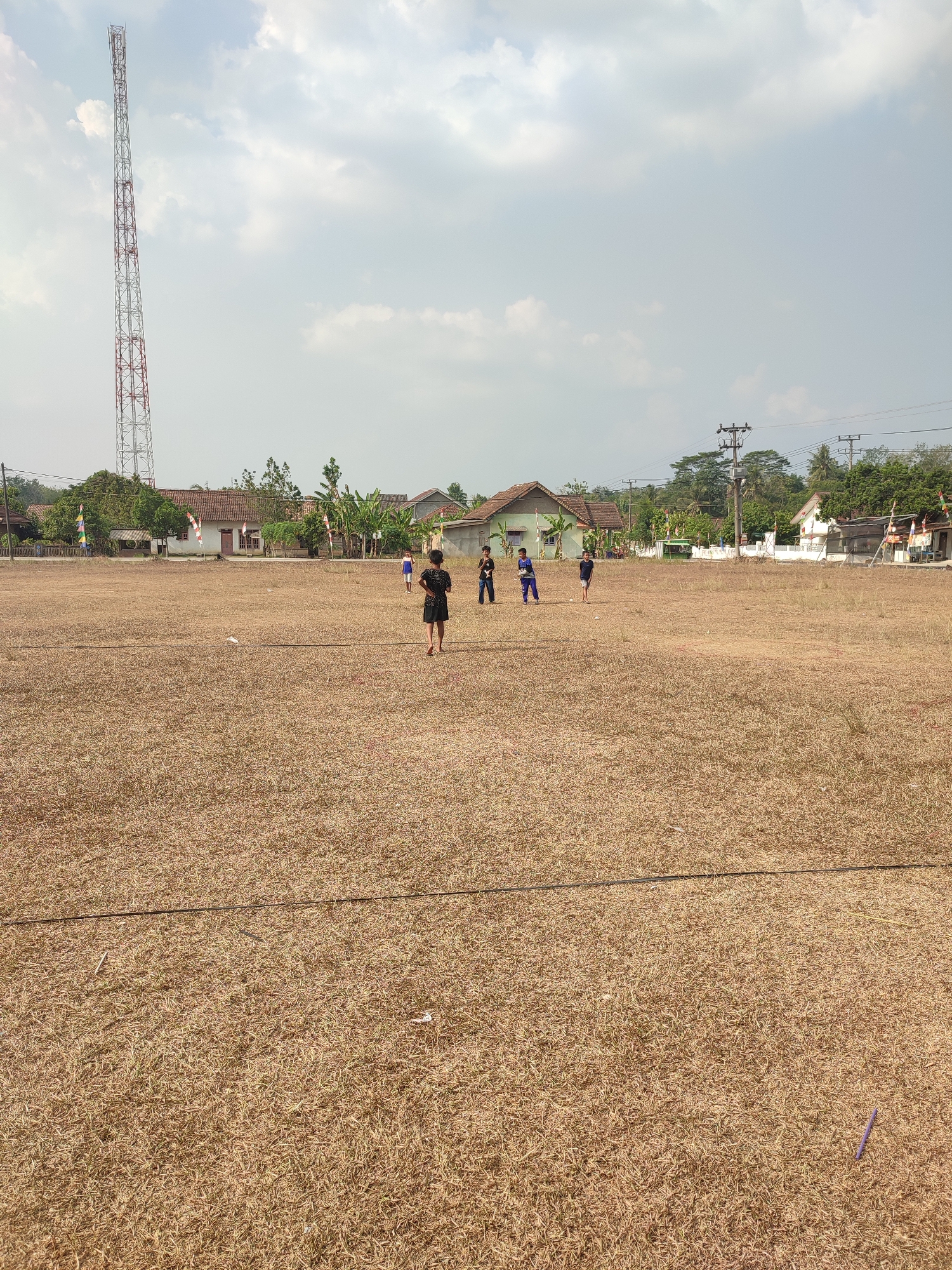 The image shows a wide, dry, grassy field in Nusa Jaya, Indonesia, under a partly cloudy sky. In the distance, a tall telecommunications tower stands prominently on the left side of the frame, with a cluster of houses and trees behind it. Several children are scattered across the field, some appearing to be playing. One boy in the foreground is wearing a black t-shirt and shorts, walking away from the camera. Other children are visible further back, dressed in casual clothing. The field is predominantly dry and brown, with some sparse green patches. Power lines and poles are visible in the background, suggesting the area is residential or semi-rural. The overall scene depicts a casual outdoor setting with a focus on the open field and the distant structures.