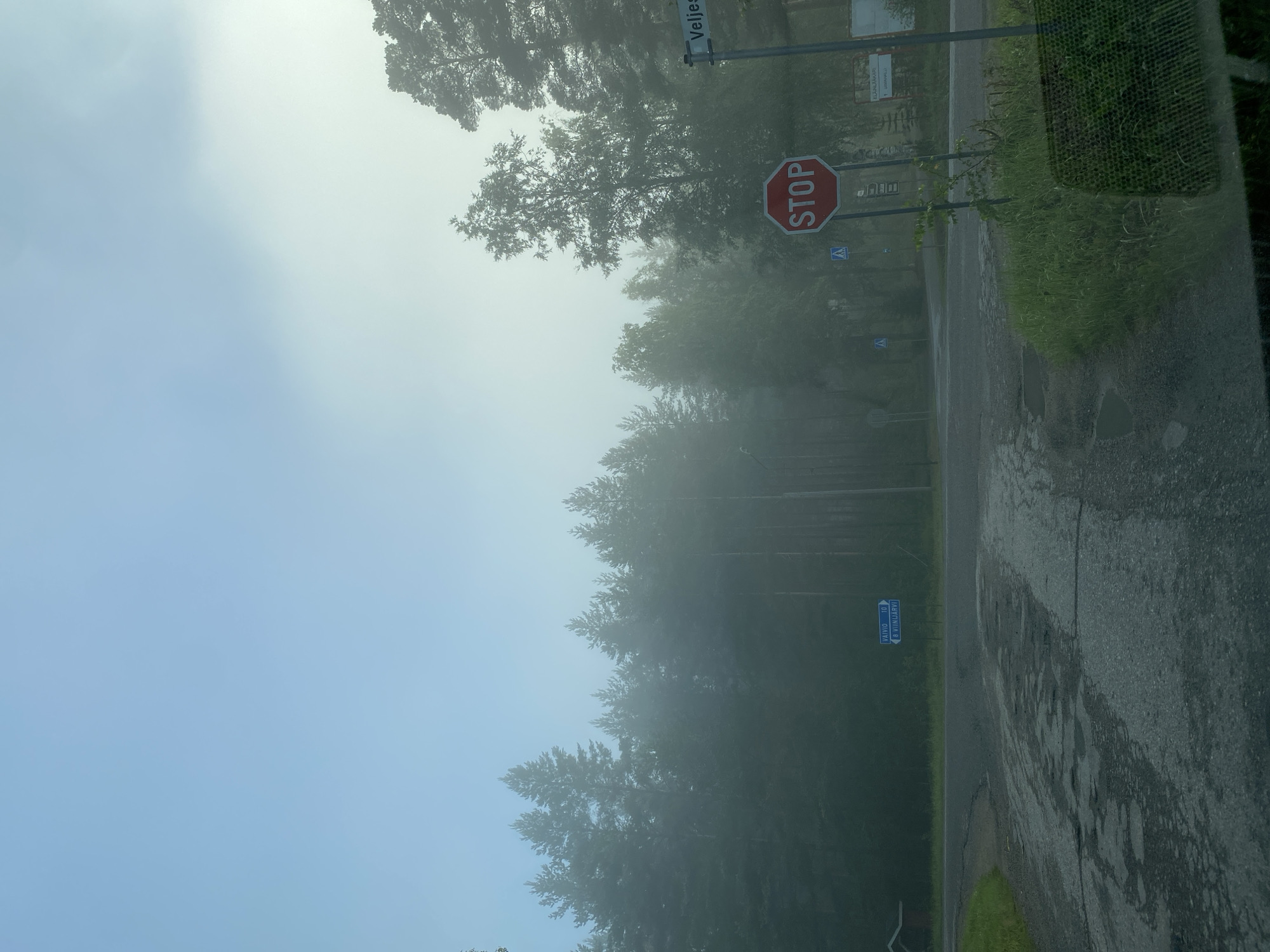 The image depicts a foggy intersection in Liperi, Finland. A stop sign is prominently displayed, along with pedestrian crossing signs and directional signs indicating "VAIVIO 10" and "P. VIINUARVI." The road appears wet and weathered. Dense coniferous trees line the background, fading into the fog. A small red building is visible to the left. The overall atmosphere is serene and slightly overcast due to the fog.