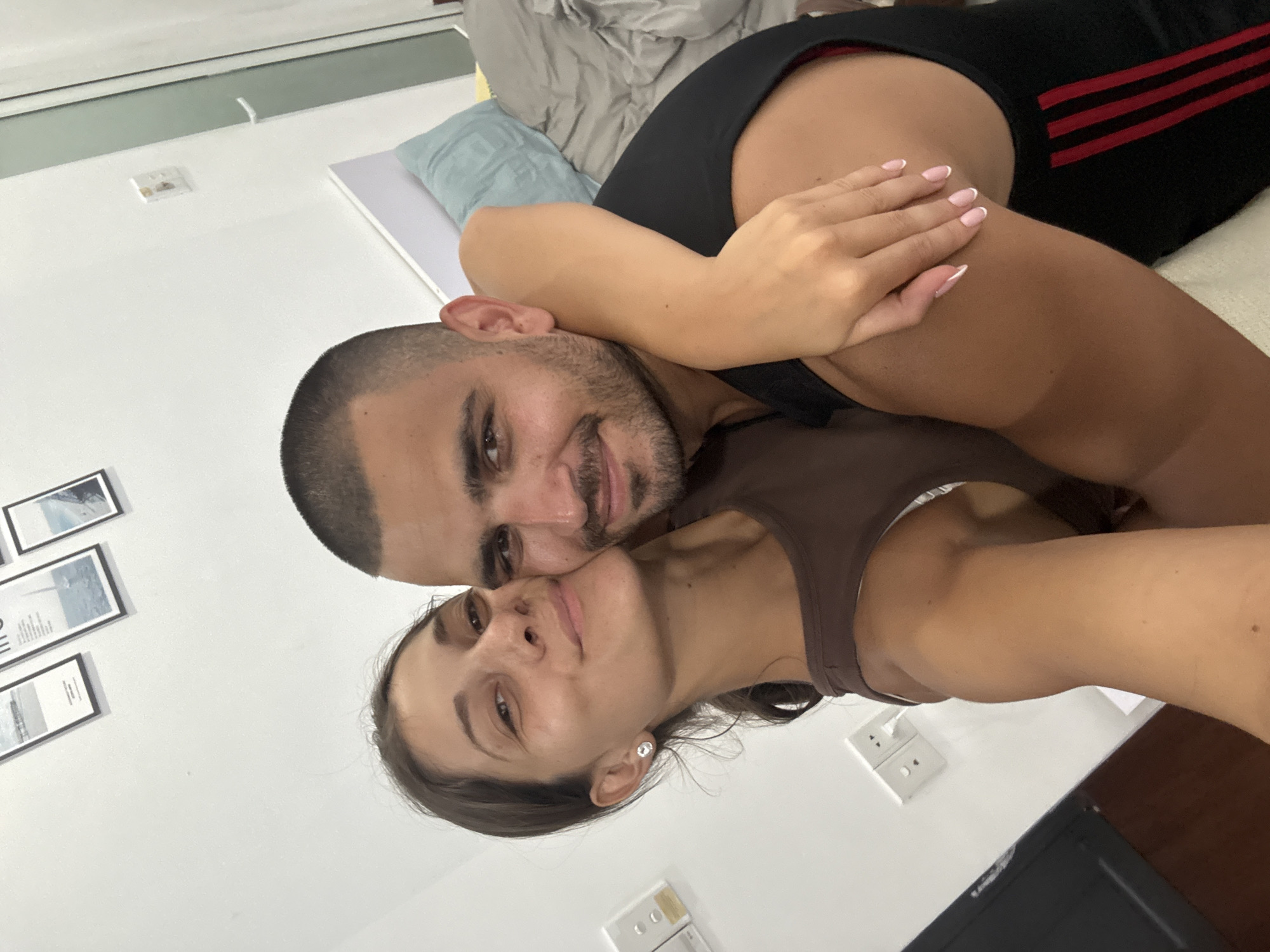 A selfie captures a smiling couple in a cozy indoor setting, possibly a bedroom or living room, with the camera looking down on them. The scene is bright, suggesting daytime. The man, with short dark hair and a beard, lies behind the woman, looking up at the camera with a gentle smile. He wears a dark top, and a black garment with red stripes (possibly Adidas) is visible behind him.
The woman, with dark hair and a small, sparkling earring in her left ear, rests her head close to the man, also looking up and smiling subtly. She wears a dark brown tank top, and her hand, featuring a French manicure, is gently placed on the man's arm. Both subjects convey warmth, happiness, and intimacy.
The background features a plain white wall adorned with a vertical arrangement of three framed pictures displaying minimalist or abstract art. Several white electrical outlets, typical of the region, are visible on the wall. A light blue pillow is seen near the man's head, suggesting they are lying on a bed or a soft surface. Part of a window or blind structure is visible in the upper left, and a dark screen, likely a television, appears in the lower right. The setting hints at a modern residence in Đà Nẵng, Vietnam.