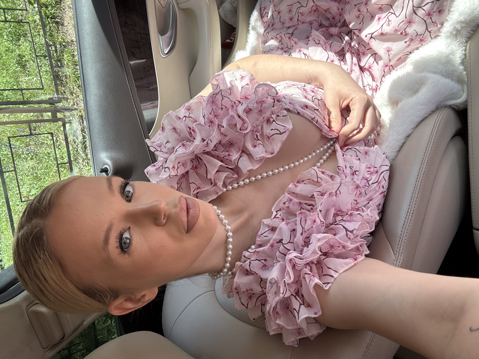 The image shows a blonde woman with her hair pulled back sitting in the passenger seat of a car. She is wearing a pink floral dress with ruffled sleeves and a pearl necklace. Her left hand is holding the pearls near her chest. The interior of the car appears to be leather and light-colored. Outside the car window, greenery and a fence are visible, suggesting a location with vegetation. The natural lighting indicates it is daytime. The image does not provide any specific indicators to place the scene in Saint Petersburg, Russia.