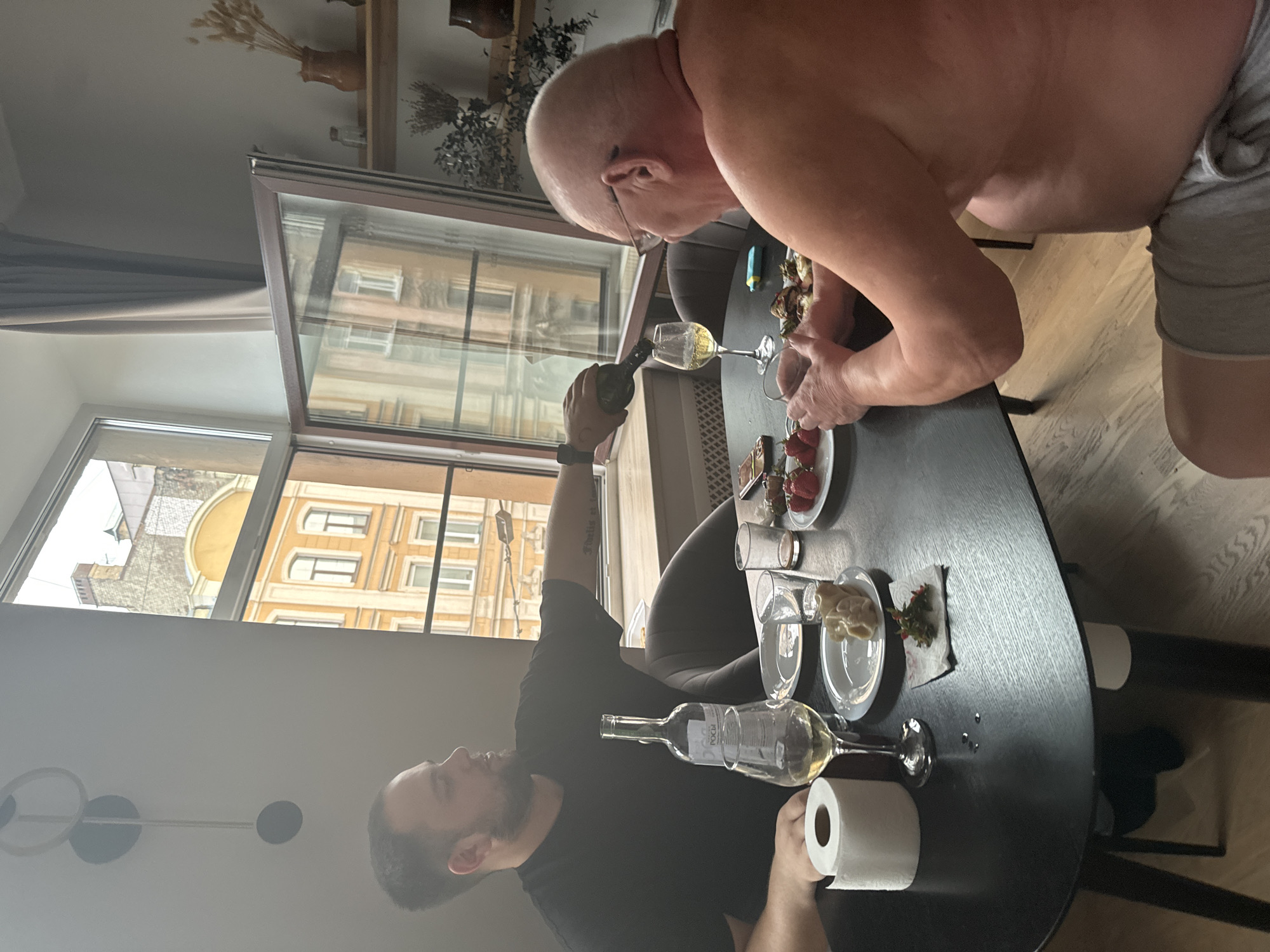 The image depicts two men sitting at a black, oval dining table. The man on the left, with a beard and wearing a black t-shirt, is pouring white wine from a dark bottle into a wine glass. He is positioned to the left of the table, facing away from the camera. The man on the right, who is bald and wearing glasses and grey shorts, is facing the table, with his back to the camera. He is holding a glass of white wine.
On the table, there are two plates, one with strawberries, another with some food that appears to be dumplings, and a third small plate with a garnish. There are also glasses, cutlery, a roll of toilet paper, and a bottle of wine. The scene is set indoors, near a large window that offers a view of buildings in Saint Petersburg, Russia, characterized by their classic architectural style with yellow facades and numerous windows. The light suggests it is daytime. A curtain is visible on the right side of the window. Shelves with decorative items are mounted on the wall to the right. The floor is made of light-colored wood planks.