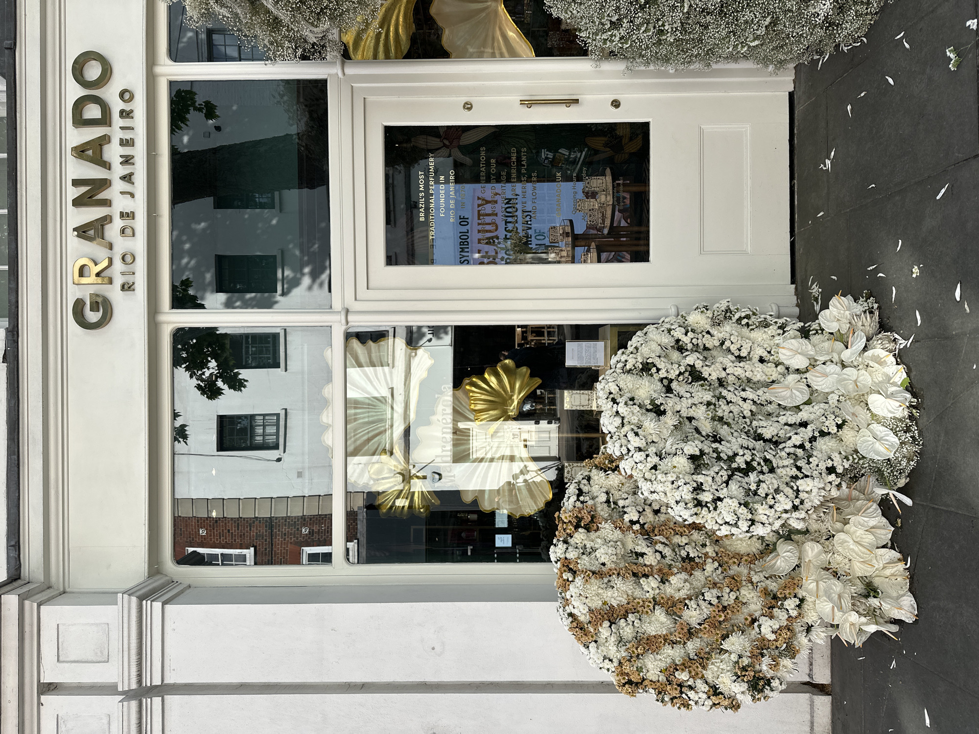 A street-level view of the front facade of a Granado store, Rio de Janeiro, as indicated on the sign above the storefront. The shopfront is white and decorated with floral displays. The sign “Granado” is in metallic gold. A door is visible with a sign that includes the text "BRAZIL'S MOST TRADITIONAL PERFUMERY FOUNDED IN RIO DE JANEIRO IN 1870” and "SYMBOL OF BEAUTY", and "VAST HERITAGE". Two large, round floral arrangements lie on the ground in front of the shop. The floral displays are predominantly white, with touches of brown. The location is likely in London, United Kingdom. The photo captures a quiet urban scene.