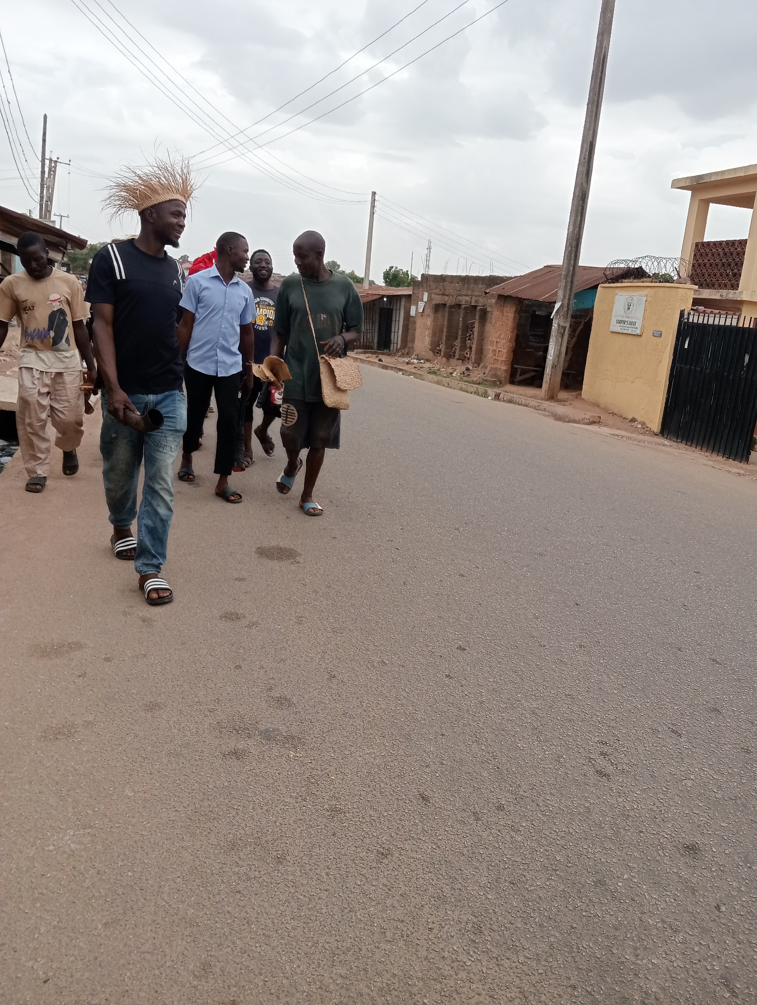 A group of six men are walking along a street in Jos, Nigeria. The man in the front is wearing a hat made of straw and carrying a horn. The next man is wearing jeans, and the third man is wearing a light blue shirt. The fourth man is wearing a t-shirt with a logo on it. The fifth man is bald. The last man is wearing a green shirt, shorts, and is carrying a straw bag.
The street is paved and lined with buildings. There are overhead power lines. The sky is overcast. A sign on the building to the right reads "Cooper's Lodge."
