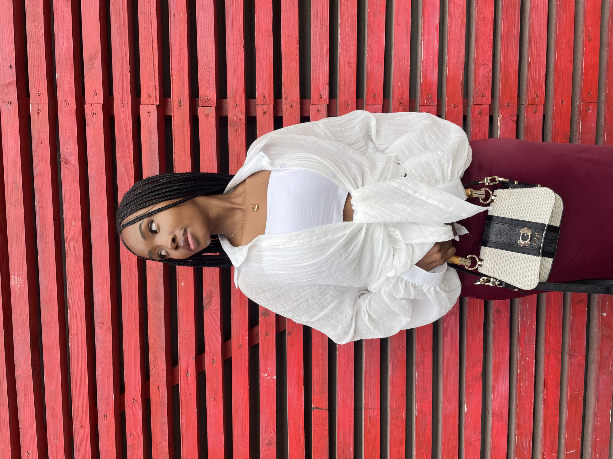 A young Black woman with braided hair stands in front of a red wooden slat wall. She is wearing a white tube top, a white loose-fitting shirt tied at the waist, and a maroon skirt. She holds a handbag with a bamboo handle and a strap. Her gaze is directed to her left. The scene is brightly lit. The image does not contain any discernible landmarks or features that would indicate it was taken in Kazan, Russia.