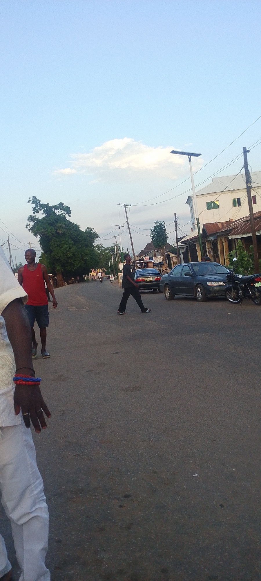 The image depicts a street in Lokoja, Nigeria, during daylight. A man in a red tank top and shorts is walking on the left side of the road, while another man in dark clothing crosses the street in the middle ground. A dark-colored sedan and a motorcycle are parked on the right side of the road. In the foreground, the arm and part of the torso of a person wearing white clothing are visible. The road surface is asphalt. Overhead power lines run along the street, and a solar street light is present. Buildings of varying styles and conditions line the street. Trees are scattered along the roadside.