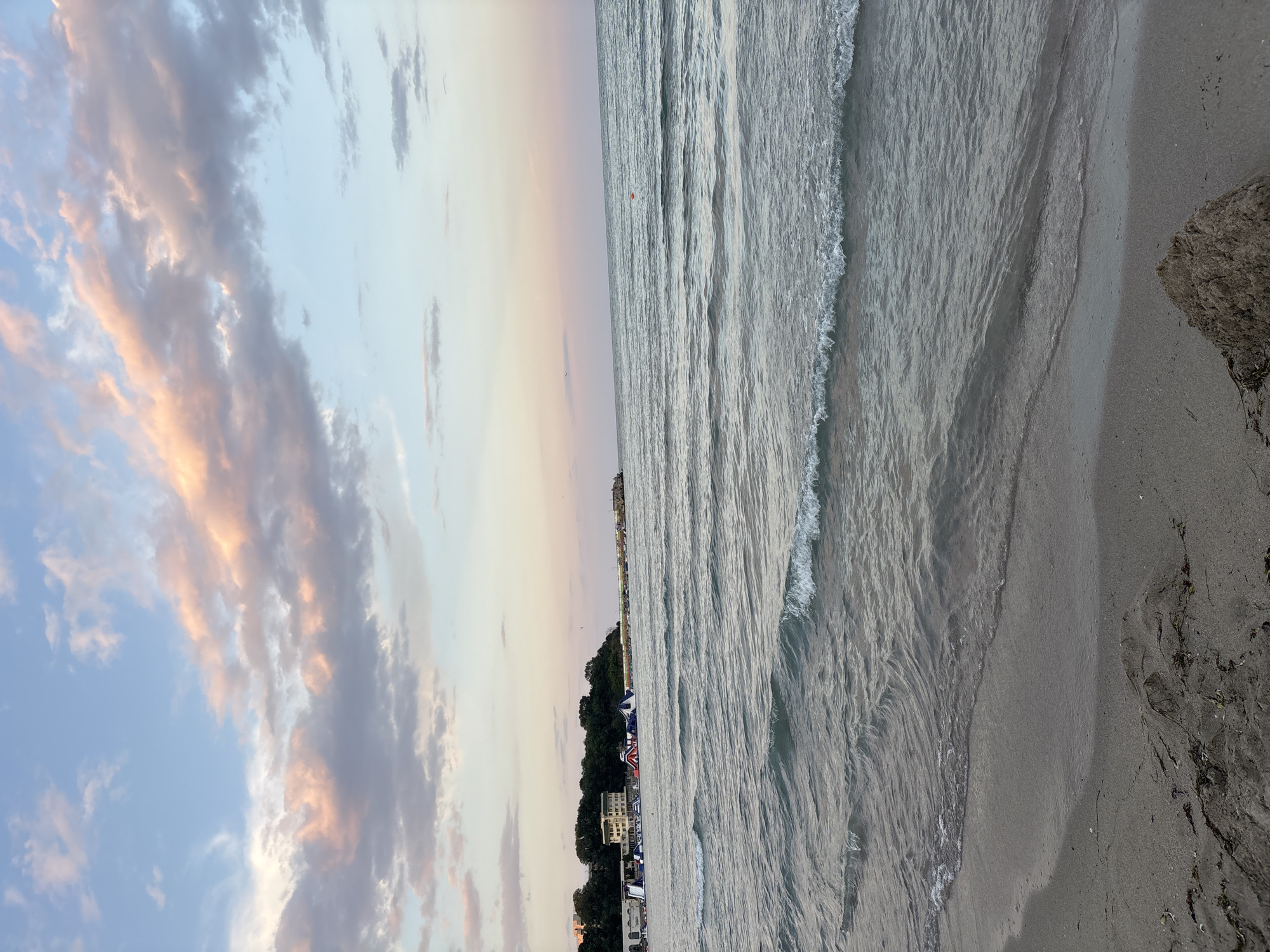 This photograph depicts a serene beach scene in Kiten, Bulgaria, captured during the late afternoon or early evening. The sky is a blend of soft blue and pastel pinks, with scattered clouds tinged with the warm hues of the setting sun. The sea is calm, with gentle waves lapping at the sandy shore.
In the distance, along the coastline, a cluster of buildings and lush green trees are visible. There are also inflatable water attractions set up on the water, suggesting a recreational area. The beach itself appears to be sparsely populated, with no prominent individuals or specific actions being depicted. The foreground shows the sandy texture of the beach, with some scattered seaweed and a small pile of sand. The overall atmosphere is peaceful and tranquil, characteristic of a seaside resort town.