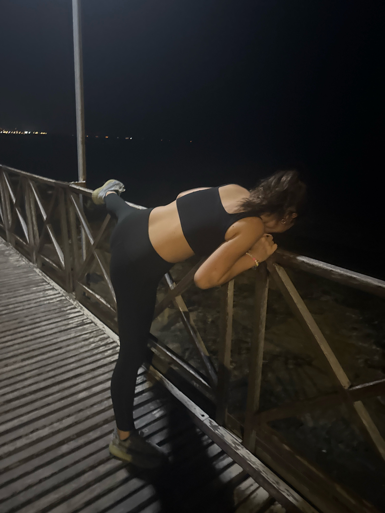 A young woman is captured in a dynamic pose at night on a wooden pier in Agios Tychona Community, Cyprus. She is wearing black leggings and a black sports bra, her right leg extended upward and resting on the wooden railing of the pier. Her left leg is bent at the knee, supporting her weight. Her body is angled towards the railing, with her arms resting on it. Her hair is dark and curly, pulled up. The pier's wooden planks create a distinct striped pattern underfoot. In the background, the dark water stretches out to meet a distant shoreline dotted with faint lights, suggesting an urban presence across the sea. A tall, slender pole, likely a light post, stands vertically on the left side of the image. The overall ambiance is dark, with artificial lighting from the distant shore and possibly unseen sources illuminating the scene.