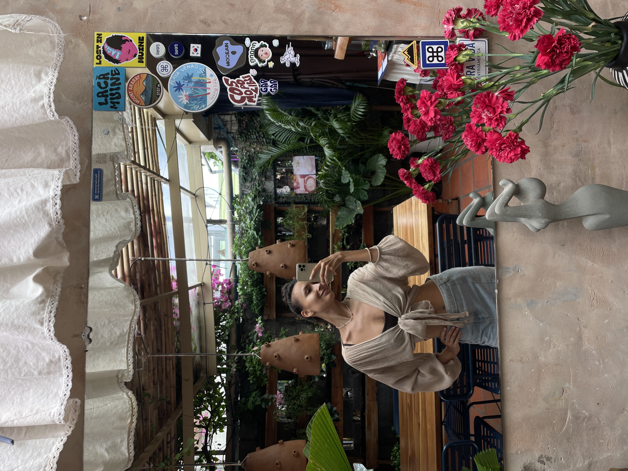 A young woman with dark hair pulled back in a bun is taking a mirror selfie in a restaurant. She is wearing a light-colored, tie-front blouse over a dark bralette and a light-wash denim skirt. The restaurant appears to be an outdoor or semi-outdoor space with a thatched roof overhead, visible through the mirror. There are wooden tables and chairs in the background, as well as lush greenery and plants. The mirror is decorated with several colorful stickers. The location is identified as Mũi Né, Vietnam. In the foreground of the mirror's reflection, a vase of bright pink carnations and a modern sculpture are visible.