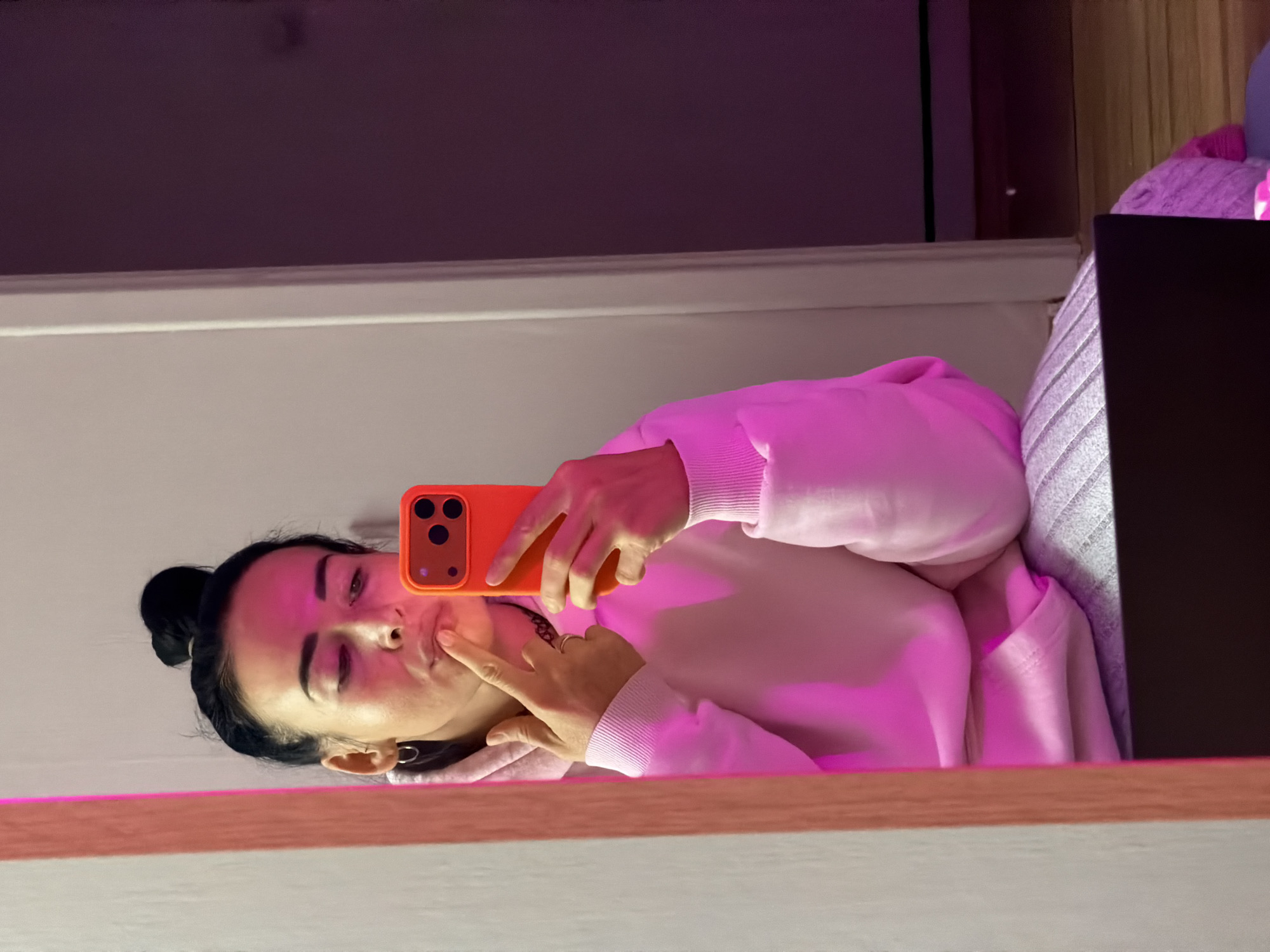 A portrait-oriented mirror selfie, rotated 90 degrees counter-clockwise, depicts a young woman in an indoor setting. The woman, with dark hair tied in a bun, is wearing a light pink hooded sweatshirt. She holds an orange smartphone with a triple camera system in her right hand, using it to capture her reflection. Her left index finger is poised near her lips, suggesting a pensive or playful expression.
The scene is bathed in a distinct pink and magenta light, casting a vibrant glow on her and her surroundings. In the reflected background, a dark wall is visible above her, and to her right, a portion of a grey patterned fabric and a dark rectangular object, possibly a laptop, can be seen. A wooden border or ledge runs across the bottom of the reflected image. The overall atmosphere suggests an evening or night setting due to the prominent artificial lighting.
No discernible text is present, and there are no specific visual cues within the image to indicate the provided location context of Saint Petersburg, Russia.