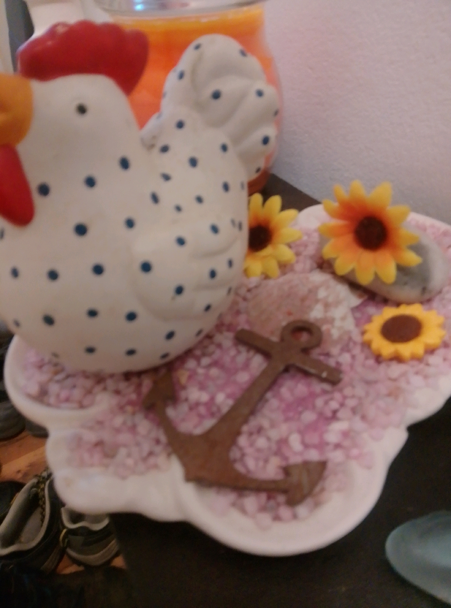 The image shows a decorative item, likely a ceramic chicken, placed on a pink, granular surface. The chicken is white with blue polka dots, and has a red comb and wattle. There are also several small decorative sunflowers and a brown anchor-shaped object on the pink surface. In the background, a blurred orange object, possibly a candle or jar, is visible. The scene appears to be indoors, with a plain white wall behind the objects and a wooden surface in the foreground. The image does not provide any information about the city of Wedel, Germany.