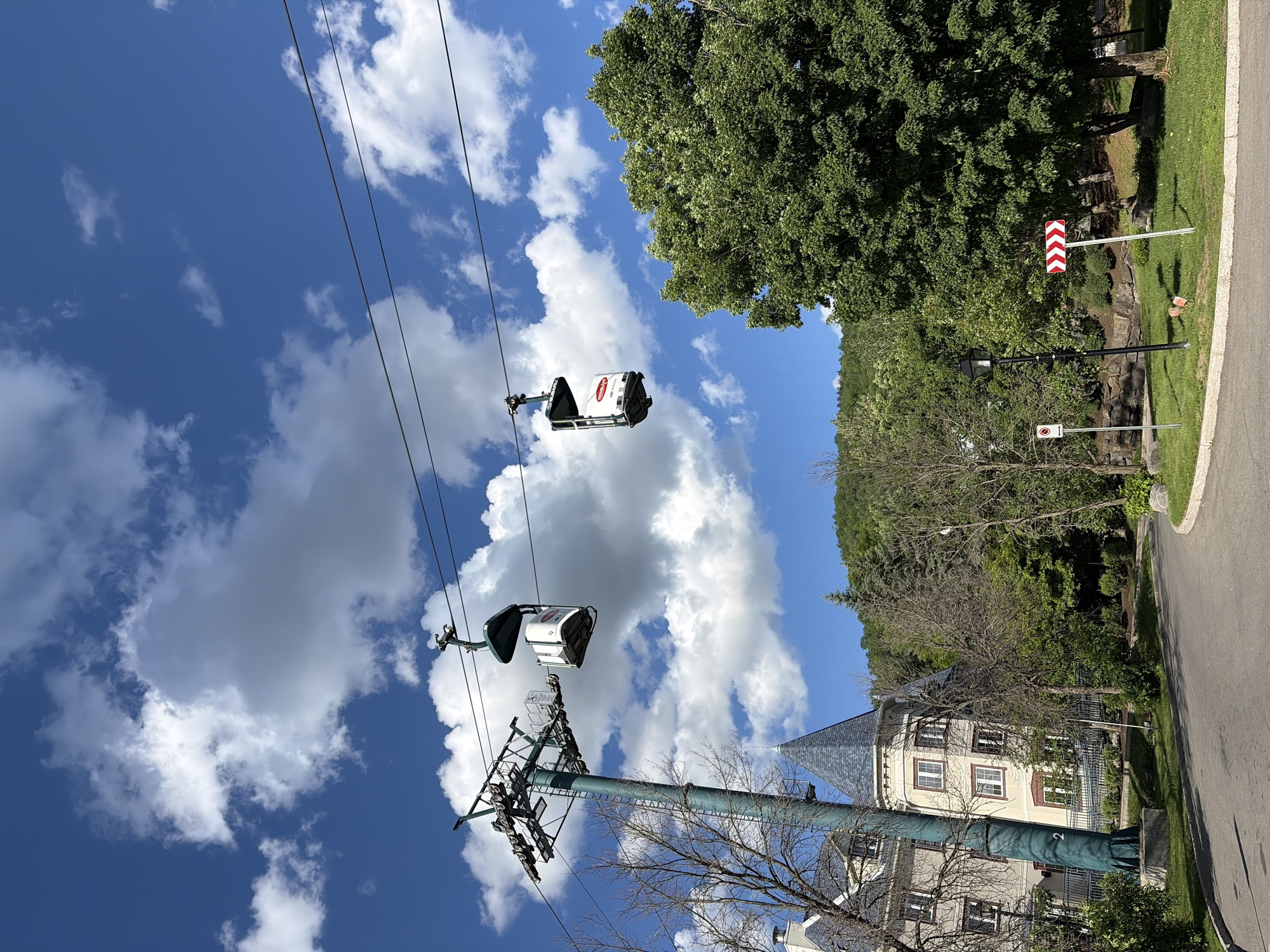The image shows a view of the Mont-Tremblant resort in Mont-Tremblant, Canada, on a clear day with scattered clouds. Two gondola cabins are suspended from cables, one on the left closer to a pylon, and another further to the right. The resort's architecture is visible in the background, with a building featuring a distinctive turret and pitched roof to the left. A paved road curves through a landscaped area with grass, trees, and a lamppost. A sign with red and white chevron arrows indicating a curve to the right is also visible. The overall scene depicts a scenic mountain resort environment.
