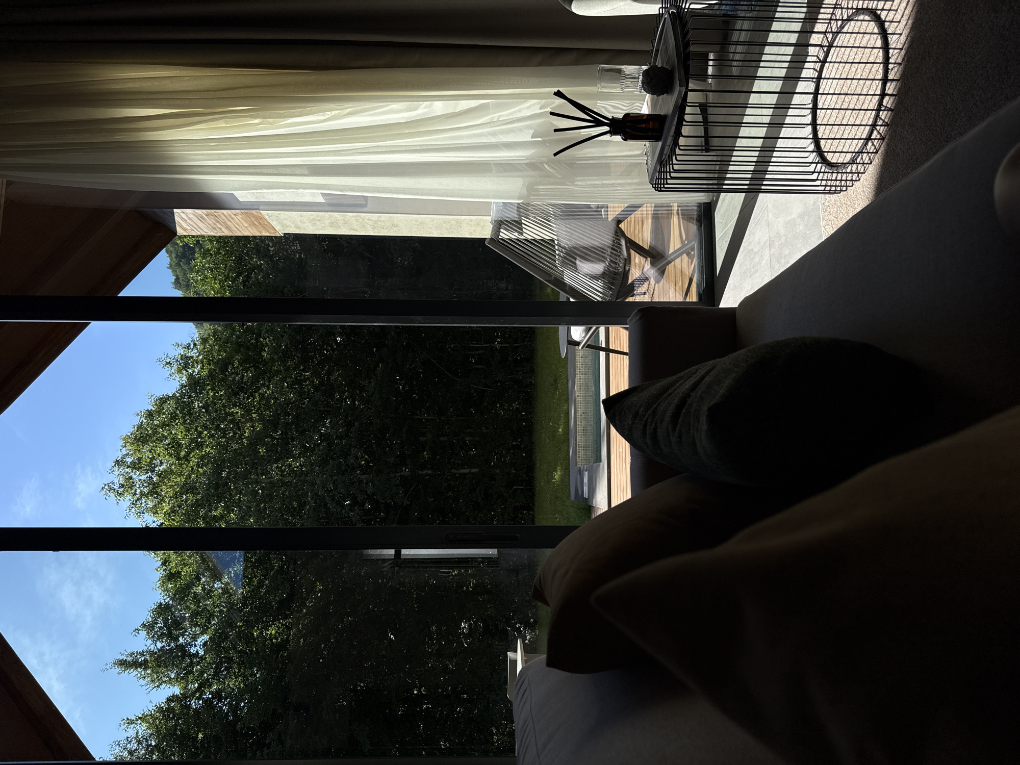 The image shows a view from a modern living space looking out through large glass doors to a natural outdoor environment. In the foreground, a portion of a sofa with dark cushions is visible, partially silhouetted by the strong sunlight streaming in. Adjacent to the sofa, a black wire side table holds a glass of water, a small decorative stone, and a diffuser with reeds in a dark bottle.
Beyond the glass doors, a wooden deck leads to a small, partially visible swimming pool. A lounge chair is placed on the deck. The outdoor area is surrounded by lush green trees and a grassy lawn. The sky is a clear, bright blue. The architecture of the building features angled wooden roof beams and concrete or stone walls. The lighting suggests it is daytime, with dramatic shadows cast by the sunlight. The overall scene evokes a sense of modern comfort integrated with a natural landscape, likely in a resort or private residence. The location is described as Bakovići, Montenegro.