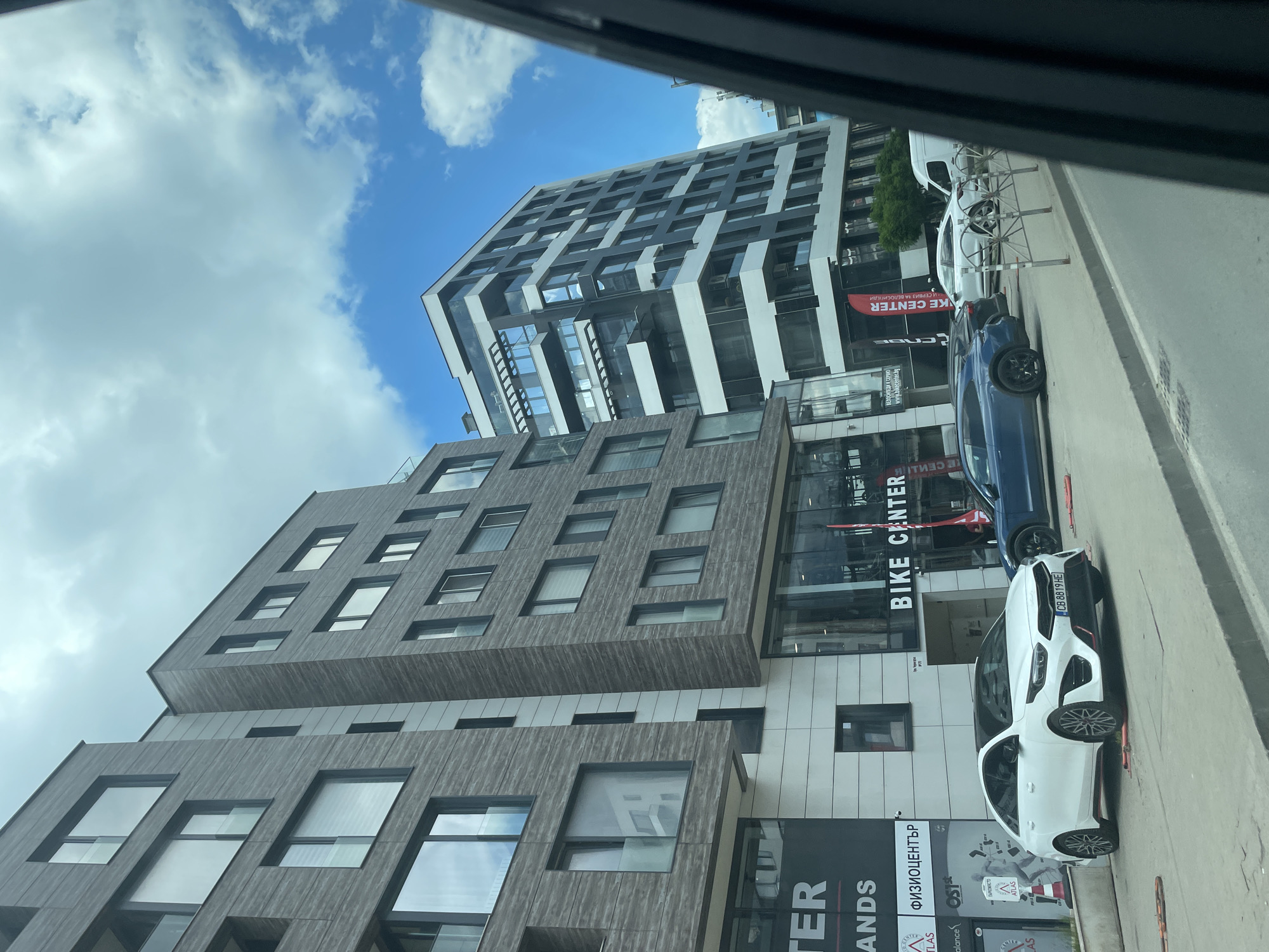 The image shows a view of modern buildings in an urban setting, likely taken from inside a vehicle. The sky is visible, with clouds and blue patches. The buildings appear to be of mixed architectural design, with one having a gray wood-like exterior and another with a modern black and white facade.
On the ground level, signs indicate a "BIKE CENTER" and a "ФИЗИОЦЕНТЪР" (Physio Center). There are cars parked along the side of the road, including a white Kia with license plate "CB 8819 HE", a blue car, and a white van. Additionally, there's street furniture visible. Based on the signs written in Cyrillic, the location is Sofia, Bulgaria.