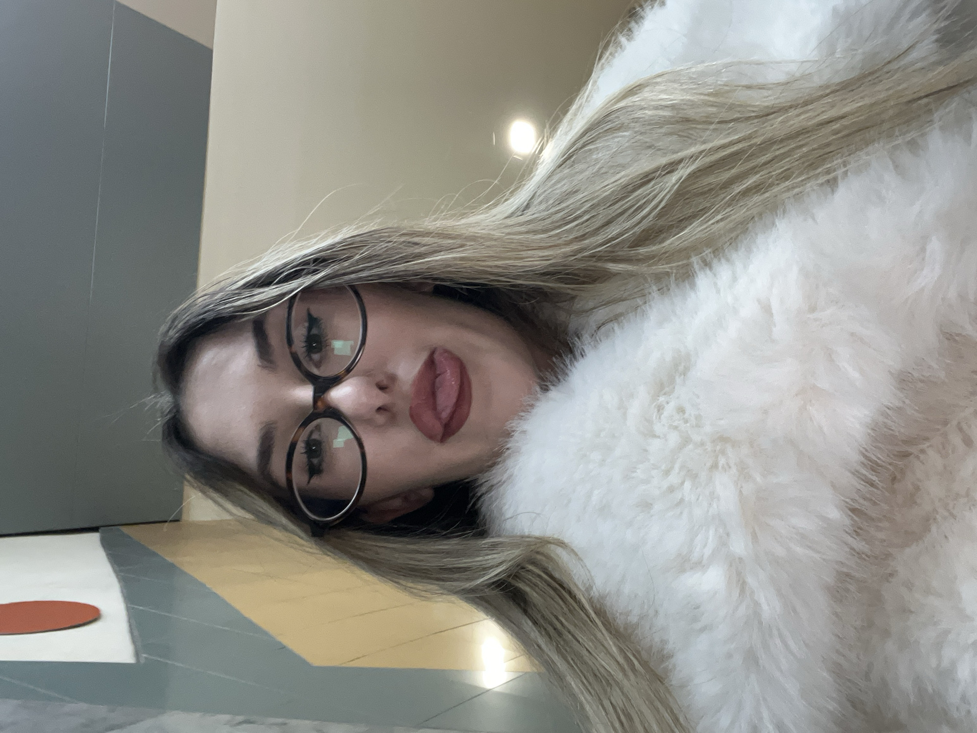 A woman with blonde hair and glasses is facing the camera, wearing a white furry jacket. The background consists of a grey wall and a beige wall. The floor is patterned with grey, yellow, and white tiles, with a red circle visible on one tile. The location appears to be indoors. The image does not provide enough information to verify that this is the city of Sofia in Bulgaria.