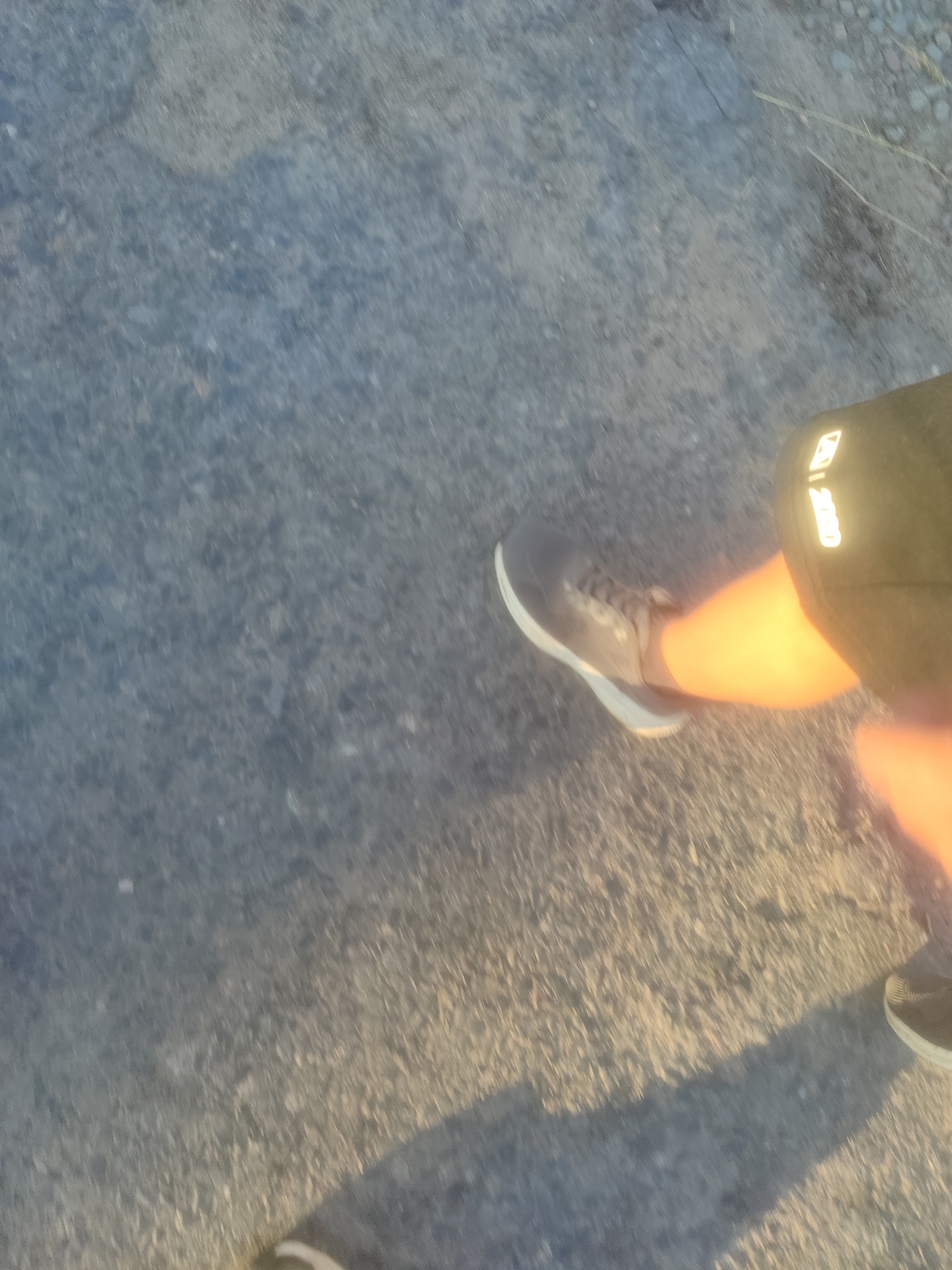 An over-the-shoulder shot captures a person's legs and feet mid-stride on a textured, grey surface, likely asphalt or concrete. The person is wearing dark shorts with a reflective logo on the right leg and a dark-colored athletic shoe on the left foot. The right leg, partially visible, is illuminated by a warm, orange light, possibly from the sun. The ground is uneven and shows some wear and tear, with small stones and debris scattered across it. The lighting suggests it is either early morning or late afternoon. The image is blurry, indicating movement.