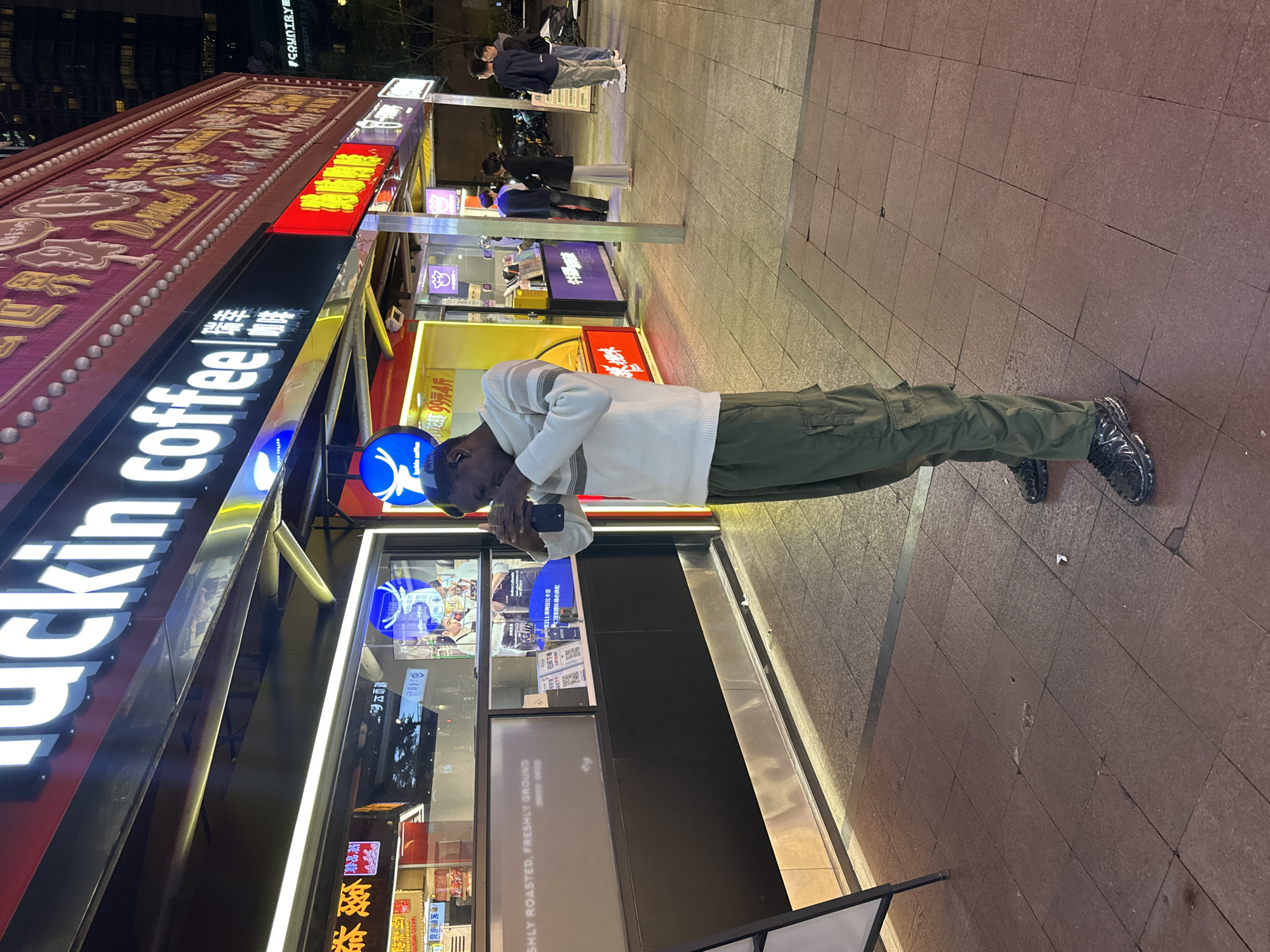 The image captures an individual standing outside a "luckin coffee" store in the Yuzhong District of China. The person is engrossed in their phone. Behind them, the store's sign is prominently displayed in English and Chinese characters. The location appears to be a bustling commercial area, as suggested by other shops and people in the background. The pavement is tiled. The time seems to be dusk or evening, as indicated by the lighting.